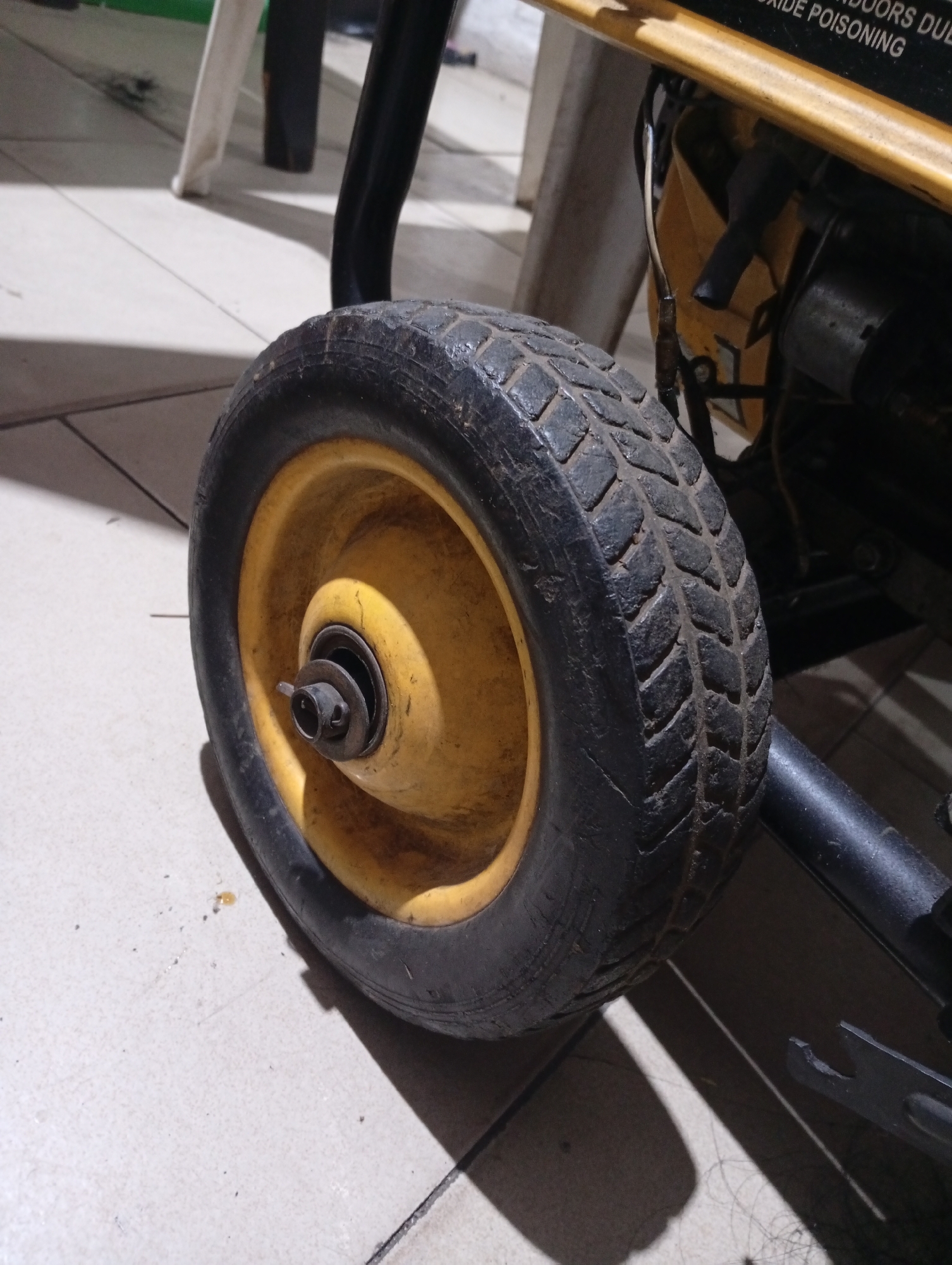 The image shows a close-up of a wheel, likely belonging to a portable generator. The tire is black and appears worn, with a yellow rim. The wheel is situated on a light-colored tiled floor. Part of the generator's frame and engine are visible behind the wheel. The scene appears to be indoors. There is no information that can be derived about the location of this image in Lokoja, Nigeria. No people, interactions, or actions are visible in the image.