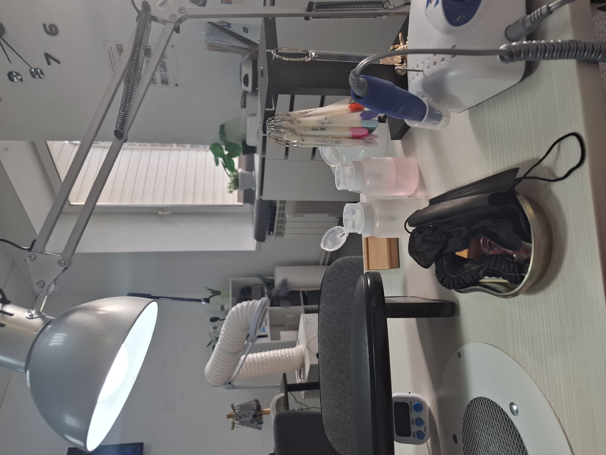 A workstation for manicures or beauty treatments is shown. The scene is indoors. An articulated lamp illuminates the workspace from the left. A white machine for filing or grinding nails is on the right. Several bottles of liquids and a small wooden block are nearby. In a metal container, there's a crumpled black face mask and black items. A timer is visible on the table's edge. In the background are white cabinets, a window, and plants. There is no evidence that confirms that the location of this image is Chaikovsky, Russia.