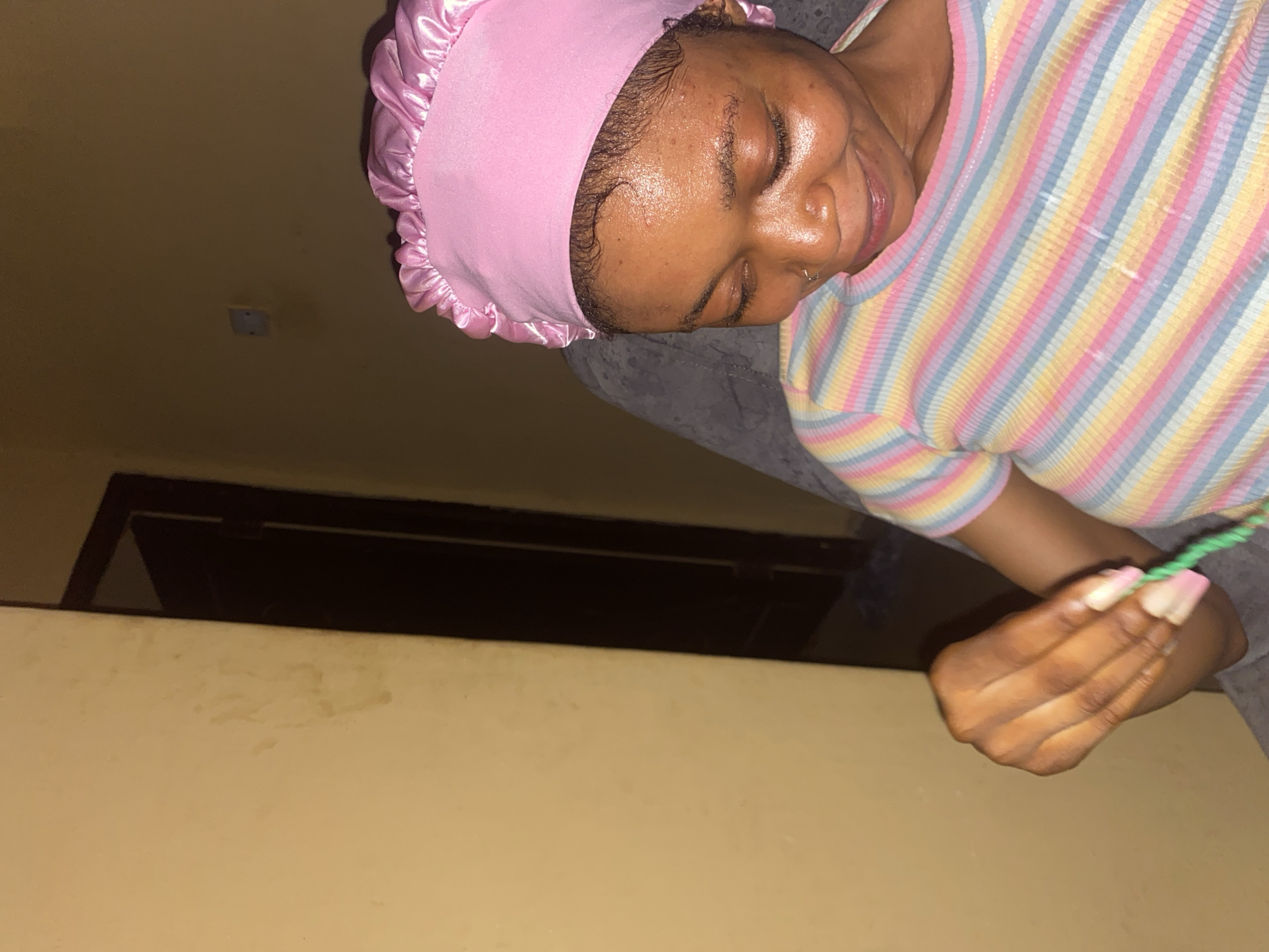 A person wearing a pink satin hair cap and a striped shirt is seated. The person is holding a green object in their right hand. The background shows a wall and a dark doorway. The location is unspecified.