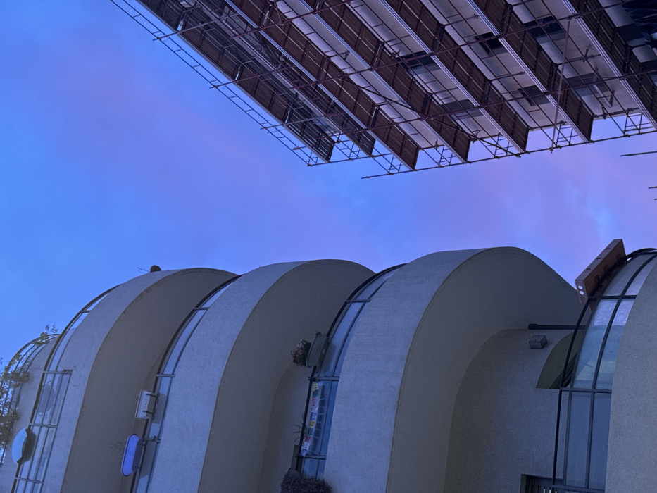 A low-angle shot reveals a modern architectural structure with rounded white walls and arched windows, against a deep blue sky. In the upper portion of the frame, a dark, grid-like structure, possibly scaffolding or solar panels, dominates the scene. The juxtaposition creates an intriguing visual contrast between organic shapes and geometric patterns under the evening sky.