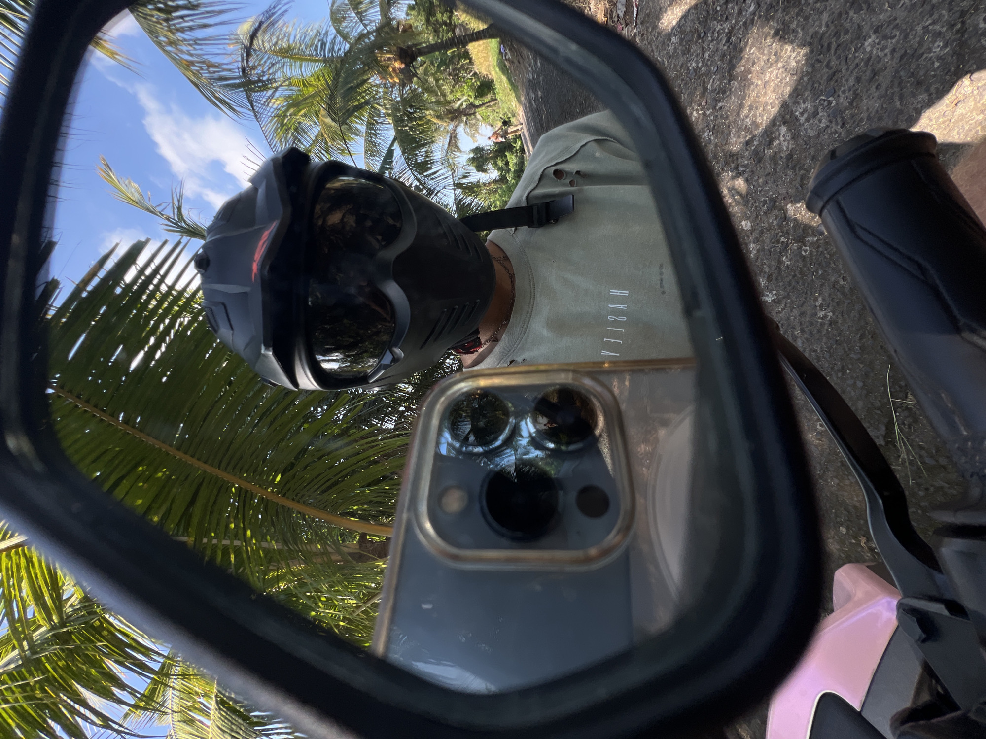 A person wearing a black motorcycle helmet with a visor is reflected in a motorcycle rearview mirror. The person is wearing a light green, ripped t-shirt with a white logo that reads "VIZHAN". In the foreground, a smartphone with a triple camera setup is reflected. The background shows lush palm trees and a bright blue sky with scattered clouds. A dirt road is visible behind the person, with another person walking in the distance. The scene appears to be in a tropical outdoor setting, possibly in Kerambitan, Indonesia, given the presence of palm trees and the overall ambiance. The ground visible in the lower part of the image is rough and rocky, suggesting a rural or unpaved road. A pink and black motorcycle handlebar is partially visible in the bottom left and right corners of the image.