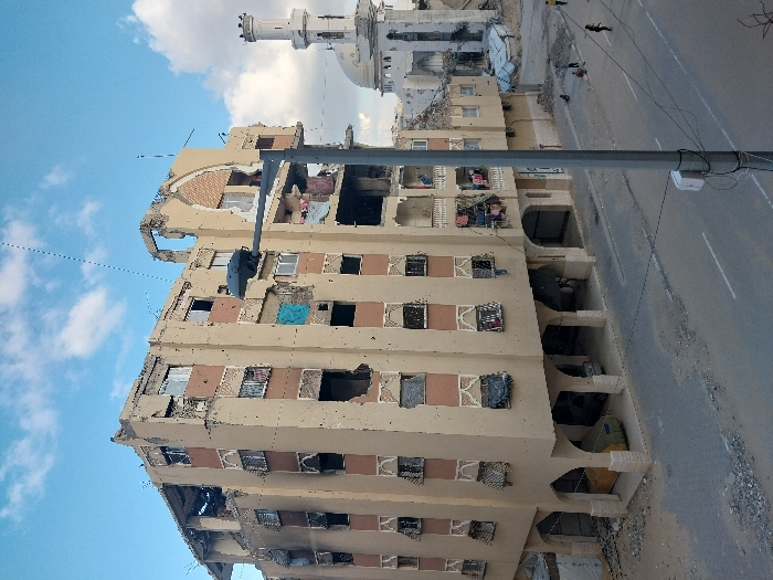 The image depicts an urban street scene in Khan Yunis, Palestinian Territory, characterized by extensive structural damage. A multi-story building, light beige with reddish-brown decorative elements, occupies the left side of the frame. It exhibits numerous broken or missing windows, facades pockmarked by impacts, and significant structural collapse on its upper floors and roof. Tarps and various fabrics cover some window openings, and laundry hangs from balconies. On the ground floor, two partially obscured vehicles are parked beneath arched supports, one covered in yellow material. To the right, a mosque with a white dome and a tall minaret stands with visible damage, including partial collapse and surrounding rubble. The asphalt street in the foreground and midground is partially strewn with debris. Several individuals are present on the street: one figure in black stands further right, another walks left, and several others are gathered near debris piles. A grey street light pole with a luminaire and attached wires stands prominently in the mid-right, partially obscuring the view. The sky above is blue with scattered white clouds.