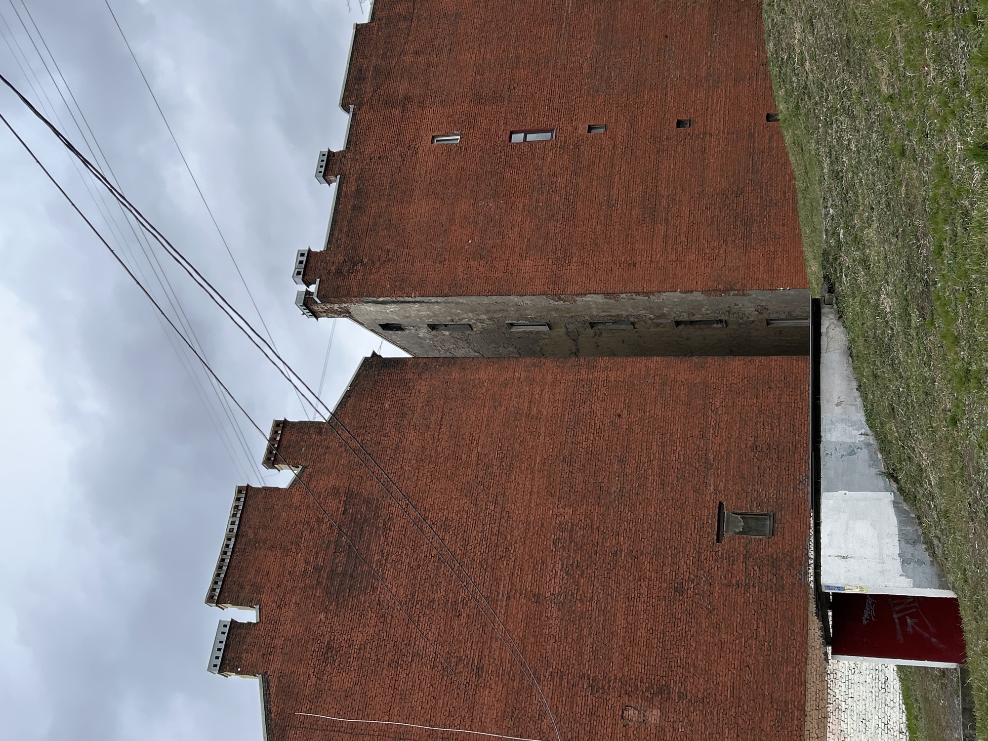 This image, captured in Saint Petersburg, Russia, depicts a narrow, upward view between two tall, imposing brick buildings under an overcast sky. The structures are built from reddish-brown brick, exhibiting signs of age and weathering. Both buildings feature distinctive crenellated or stepped gables along their rooflines. Small, rectangular windows are sparsely placed across the upper sections of the brick walls.
A shadowed, narrow passage runs between the two main buildings. The one on the right (when viewed upright) has an intermediate section of exposed concrete or stucco separating different brick levels. Adjacent to this building, a steep, green grassy embankment slopes upwards, suggesting a raised terrain feature. Multiple power lines crisscross the cloudy sky, hinting at an urban or industrial setting. At ground level, beneath the building on the left, a red surface with some illegible white graffiti is visible. The scene is static, devoid of people or specific activity, and portrays a somewhat austere architectural landscape on an overcast day.