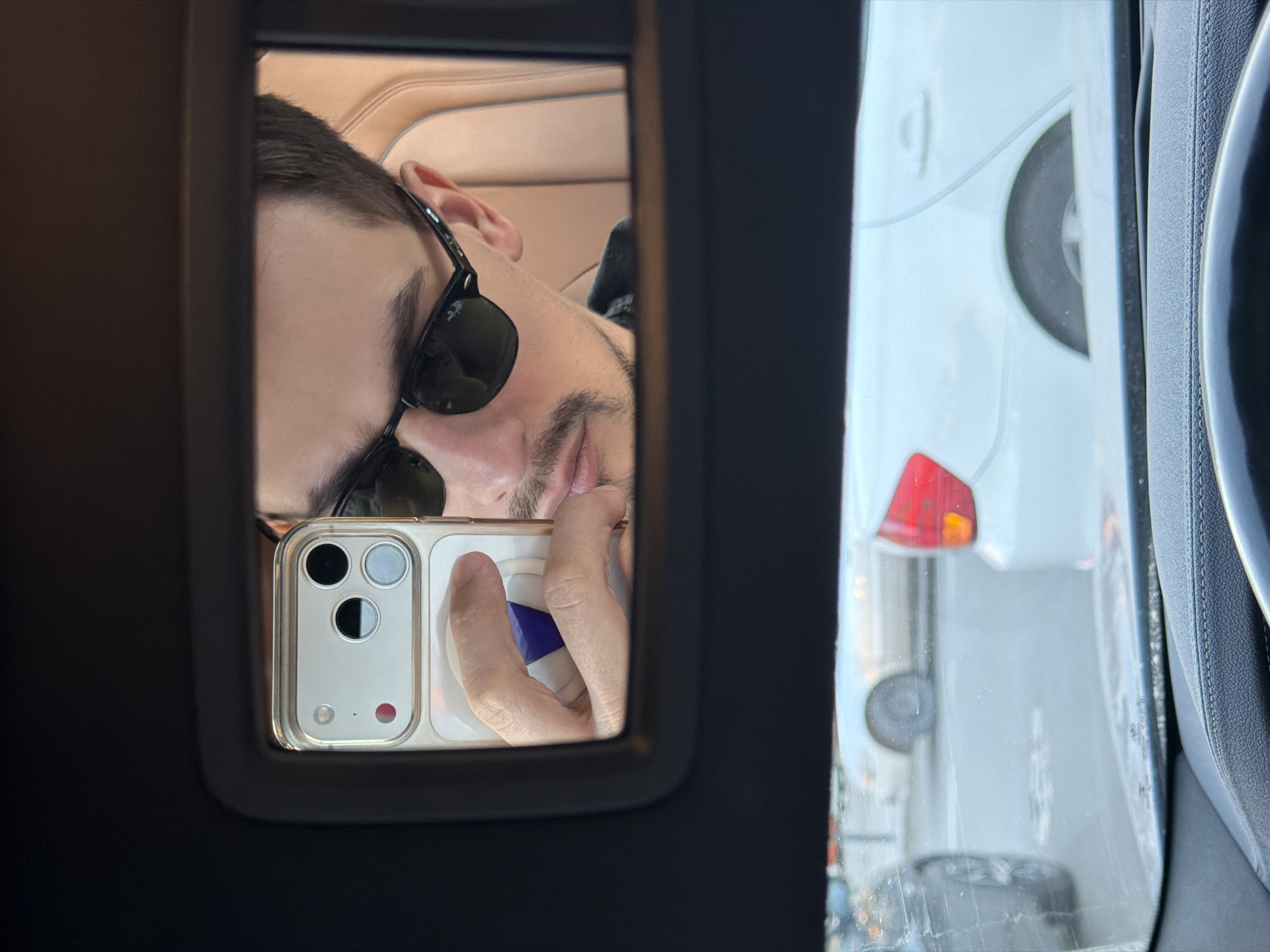 A man wearing sunglasses is taking a selfie in a car mirror. The reflection shows part of his face and his phone, with a white phone case and three camera lenses visible. Outside the car window, another white car is visible. The exact location within the city of Krasnoyarsk, Russia, cannot be determined from the image.