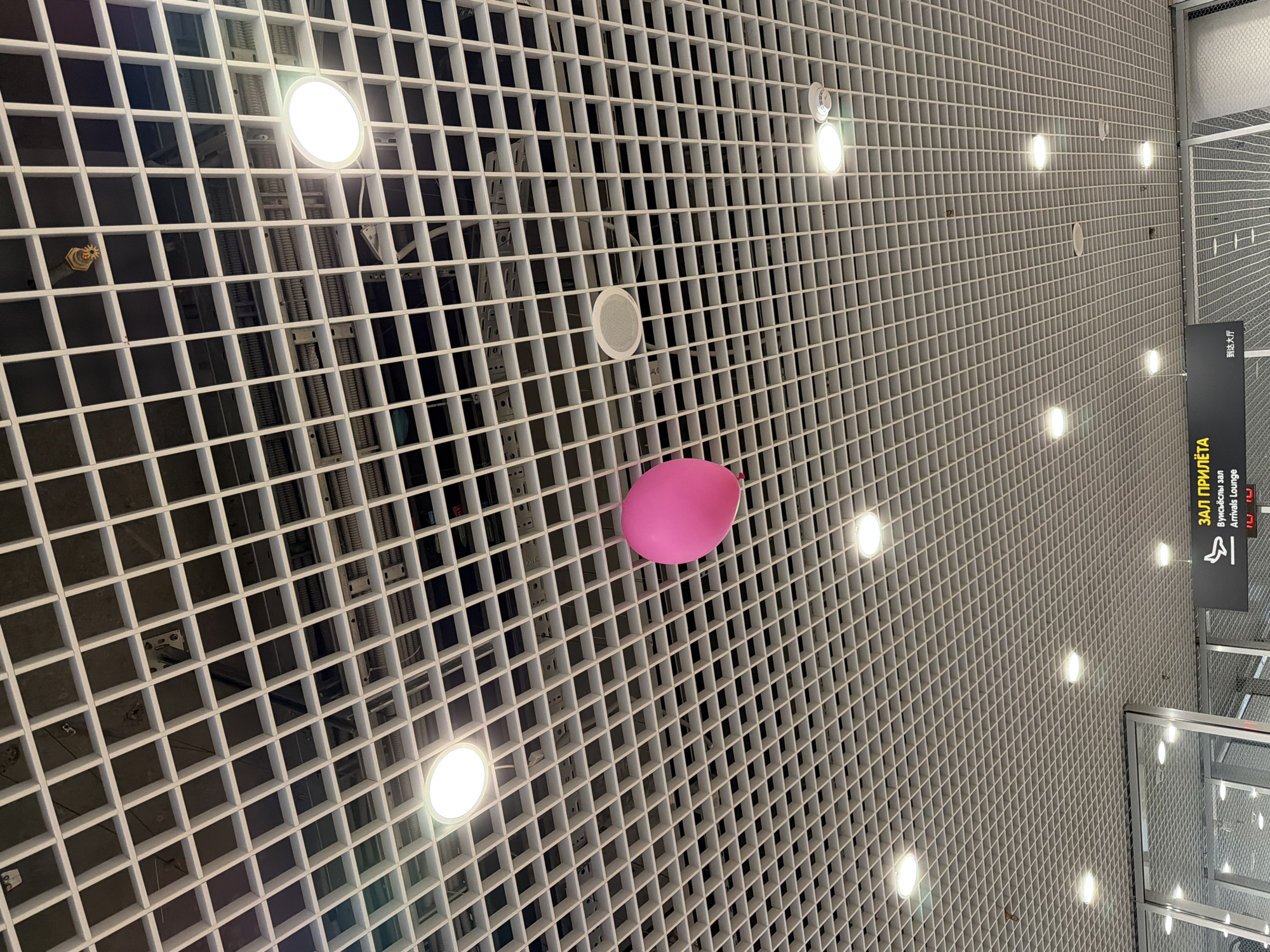 The image shows a view looking up at a square grid ceiling, likely in an airport or large public building. The ceiling is composed of small, square openings. Recessed circular lights are spaced throughout the grid, casting bright, white illumination. A single, pink balloon is suspended within the grid, appearing closer to the viewer. In the distance on the right, signage is visible with Cyrillic lettering, possibly indicating "Arrivals Lounge." The general impression is of a modern, somewhat industrial aesthetic. The location is Новое Мартьяново, Russia.