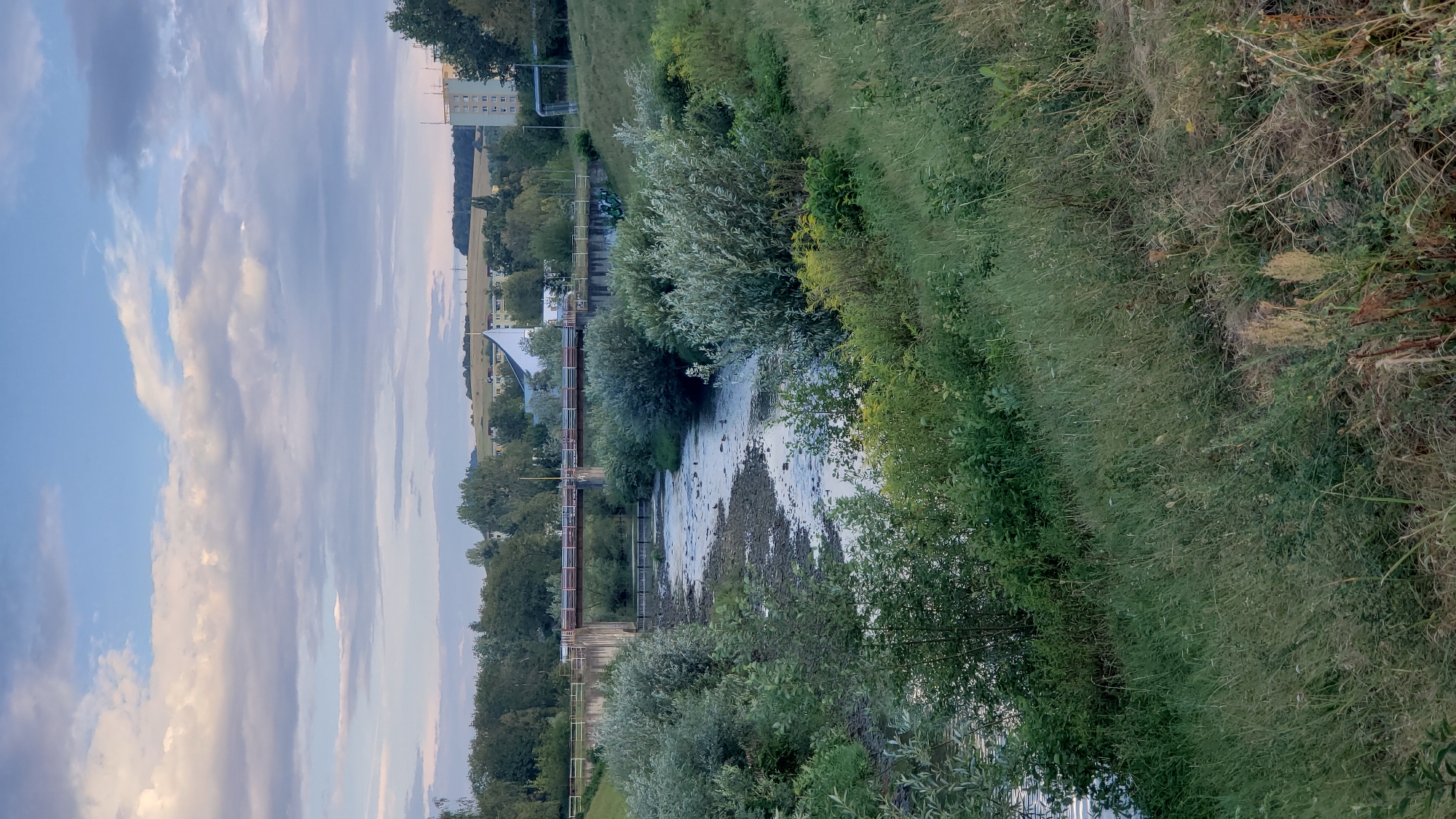 The image shows a view of the Sekčov river in Prešov, Slovakia, with a concrete bridge spanning across it. The riverbanks are lined with lush green vegetation, including trees and shrubs. In the background, there are residential buildings and a distinctive white, pyramid-shaped structure, possibly a church or a modern building. The sky above is partly cloudy, with a mix of blue sky and soft white and gray clouds. The scene appears to be taken during the daytime, likely in the late afternoon or early evening, given the soft lighting. There are no people visible in the image. The water in the river is relatively shallow, revealing rocky riverbeds in some areas, and several ducks are swimming in the water. The overall impression is a natural scene integrated with urban elements.