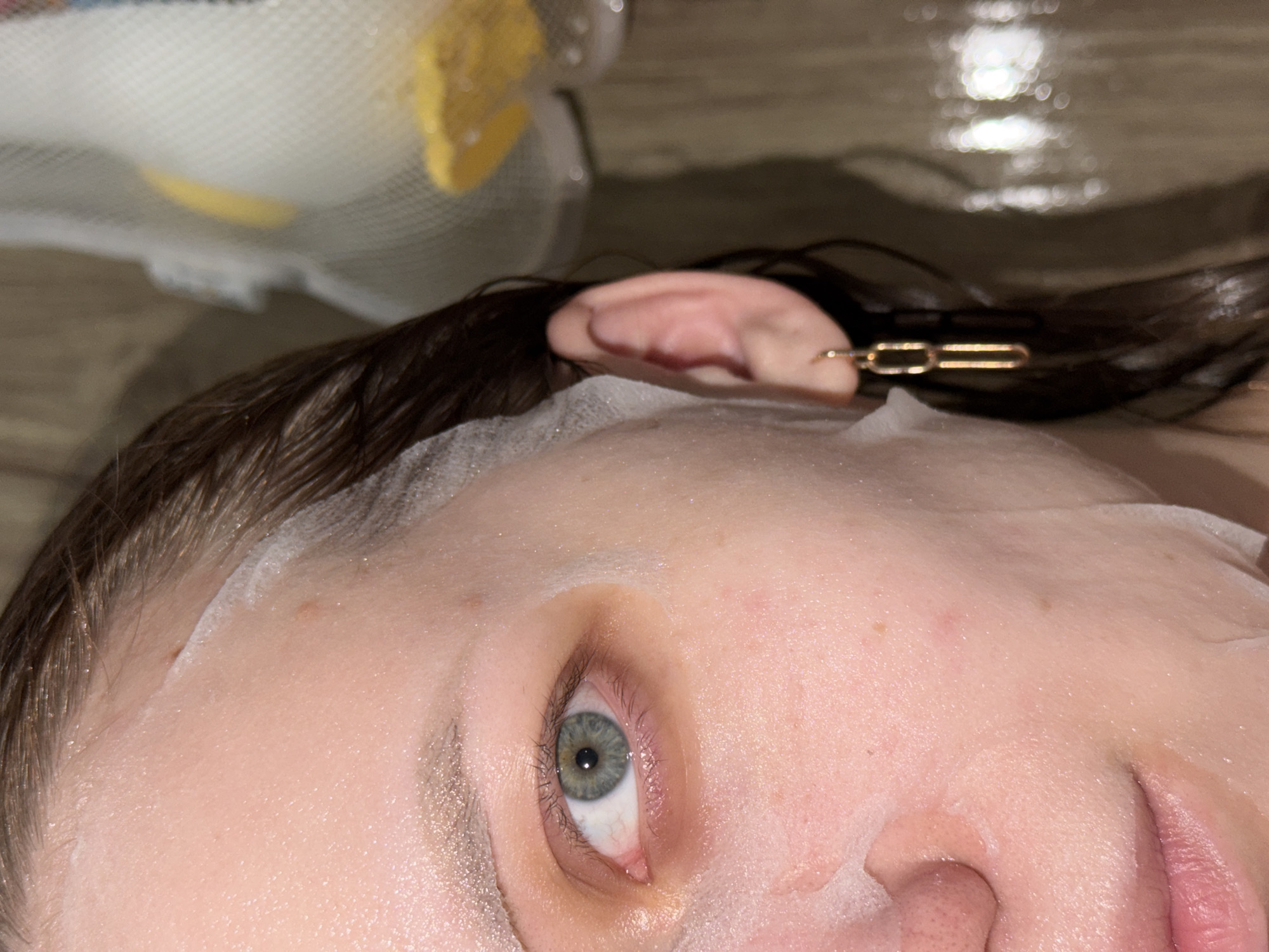 A close-up, upward-angled view focuses on a person's face undergoing a skincare treatment. A white sheet mask is applied to the forehead and cheek areas. The person's eye is visible, displaying a mix of gray and green. Their wet, dark hair is pulled back, secured with a gold-colored hair clip near the ear. A white loofah sponge with yellow accents is partially visible in the top left corner of the frame. The location is not discernible from the image.