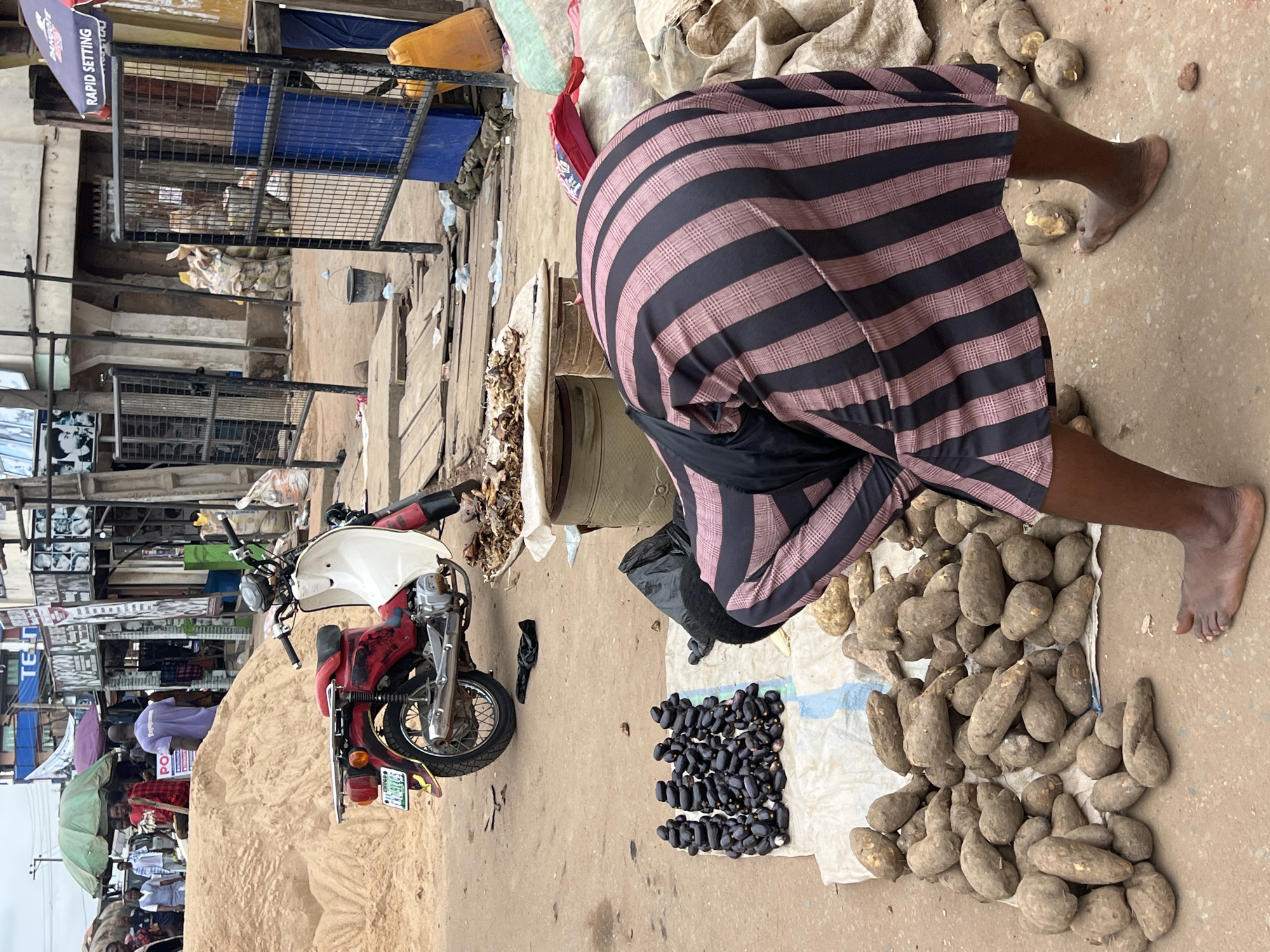 In Nkpor, Nigeria, a street market scene unfolds with a focus on agricultural produce and local commerce. A red and white motorcycle is parked on a dirt road, its license plate visible. In the foreground, a person wearing a black and pink striped garment is bent over, interacting with a pile of tubers laid out on a white cloth. To the left of the tubers, a neat arrangement of dark, elongated fruits or vegetables is displayed on a similar cloth. Behind the motorcycle, a large mound of sand suggests construction or landscaping activity. The background reveals a bustling market environment with various stalls, awnings, umbrellas providing shade, and people engaged in daily activities. Buildings line the street, some with visible signage. The overall impression is one of vibrant local trade within a busy urban setting.