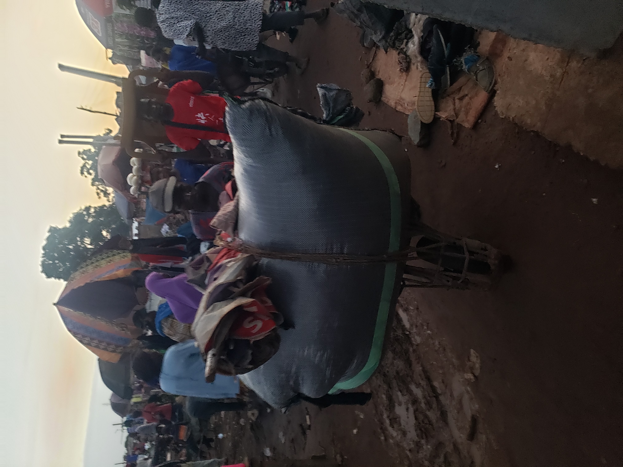A busy marketplace in Kakuri Gwari, Nigeria, is depicted at dusk. A man in a red t-shirt is the central focus, pushing a wheelbarrow loaded with a large, dark grey, cylindrical bag. The bag is secured with ropes and appears to contain bundled items. The ground is muddy and uneven, suggesting recent rain. Various market stalls with colorful umbrellas are visible in the background, along with a crowd of people engaged in their daily activities. The atmosphere is lively and bustling, typical of a Nigerian market.