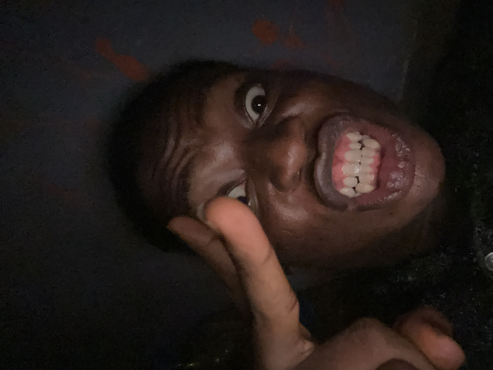 A close-up, low-angle shot depicts a dark-skinned person with their mouth open wide, displaying teeth. Their eyes are wide open, and a hand is raised with two fingers extended in what could be interpreted as a peace sign. The background is dark and unclear. The location is unknown.