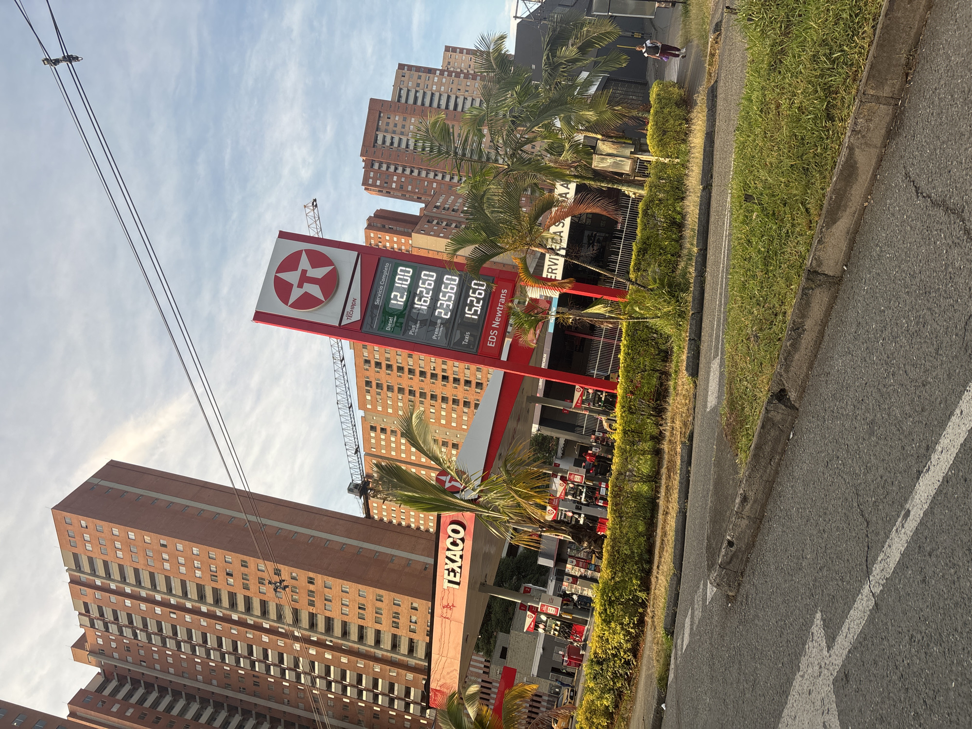 The image shows a Texaco gas station located in Bello, Colombia. The gas station has a large red and white Texaco sign with the gas prices displayed: Diesel (12.100), Plus (16.260), Premium (23.560), Taxis (15.260). "EDS Newtrans" is printed at the bottom of the price sign. Palm trees are visible next to the station's sign.
Behind the gas station, there are tall brick buildings. A construction crane is visible amongst the buildings. Wires run across the sky in the foreground. The street is asphalt with a curb and a patch of green grass separating it from the gas station. A person is seen walking on a sidewalk in the distance.