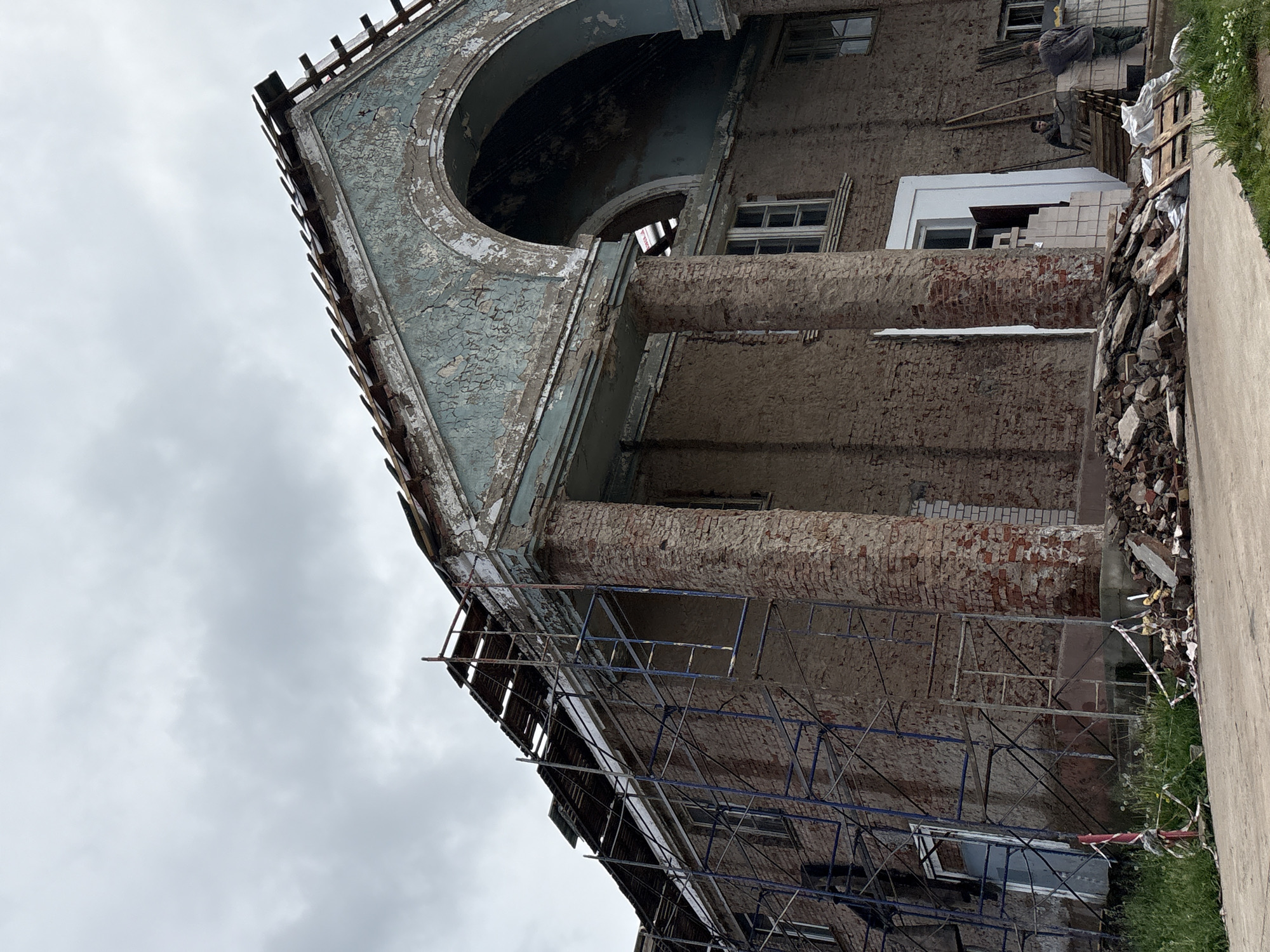 In the city of Kstinin, Russia, a large, old building is undergoing renovation. The building, constructed of brick and stucco, features a grand entrance with large pillars and an arched facade. The roof is in disrepair, with scaffolding erected around it to facilitate repairs. Debris from the renovation is piled in front of the building, including bricks, wood, and other construction materials. Two construction workers are visible near the entrance, engaged in their work. The sky above is overcast, suggesting a cloudy day. The overall scene depicts a significant construction or restoration project in progress.