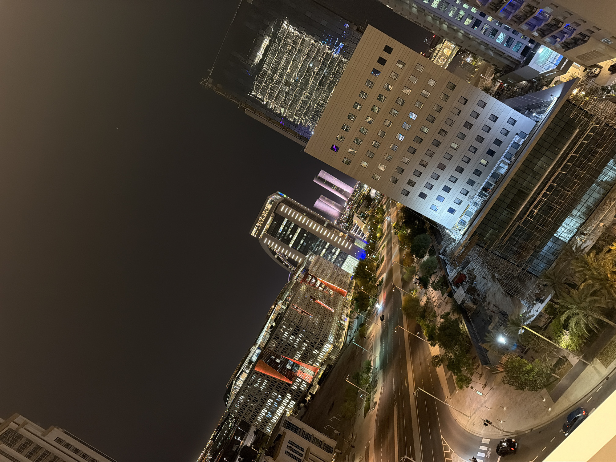 An elevated, angled night shot captures a modern cityscape with various architectural styles and illuminated buildings. In the foreground, a street with cars and lampposts winds through the scene. To the left and center, several multi-story buildings with visible windows and architectural lighting are prominent. One building features a distinctive facade with red accents. Further in the distance, more buildings, some with vertical lighting elements, are visible against the dark sky, with some appearing to be under construction with scaffolding. The overall impression is one of urban development and activity at night.