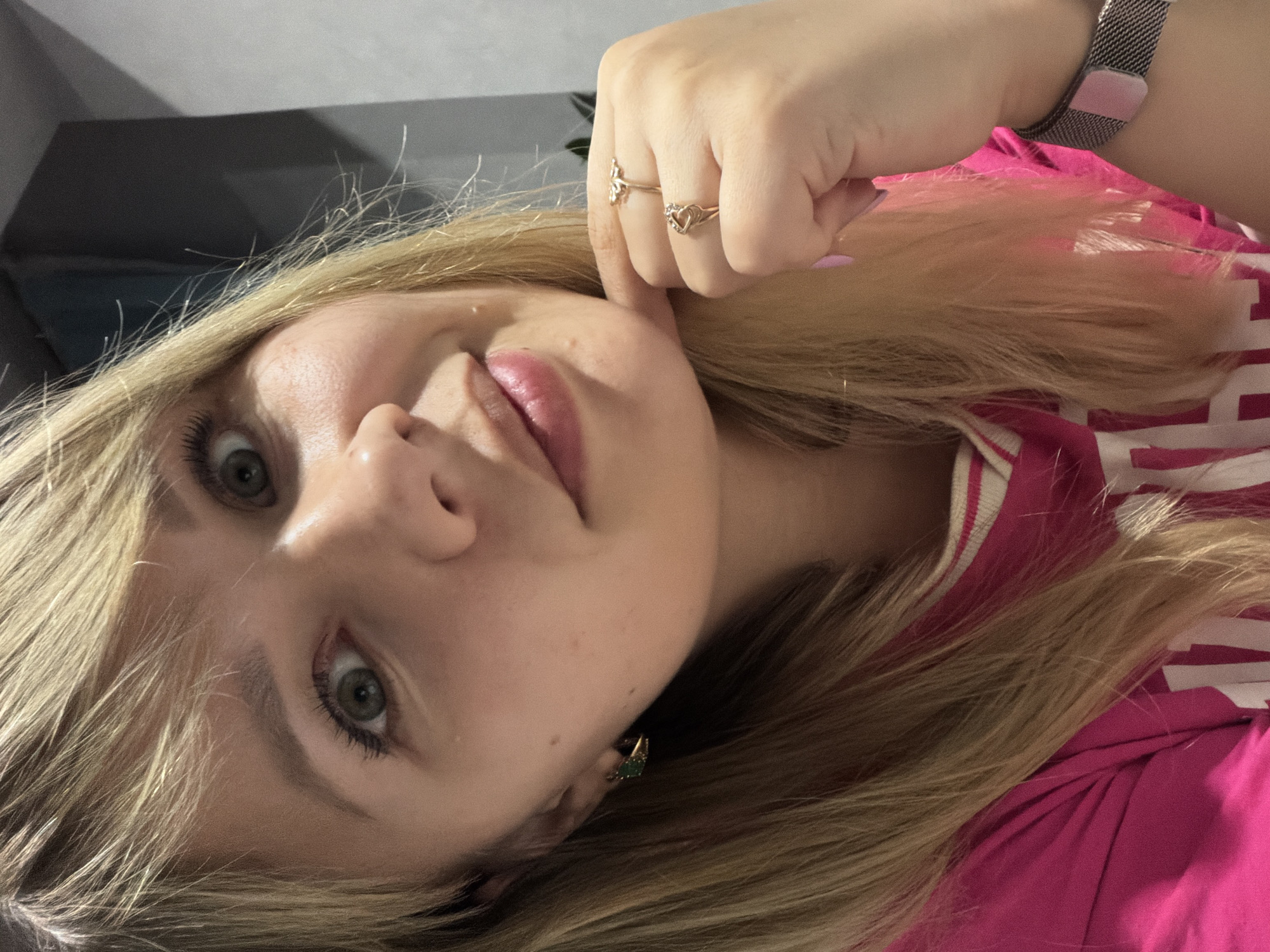 The image is a close-up, tilted portrait of a young person, likely female, with long blonde hair and green or hazel eyes. The subject looks directly at the camera with a subtle, relaxed expression, and their lips are painted a light pink. Mascara is visible on their eyelashes.
Their right hand, which has light pink nail polish, is positioned near their face, with two gold-toned rings visible on their fingers: one shaped like a butterfly, and another heart-shaped with small stones. A metallic mesh watch with a pink clasp is partially visible on their left wrist, obscured by hair and clothing. They are wearing a bright pink top, which has white stripes or a pattern on the shoulder or collar. A small, green and gold-toned earring can be seen on their left earlobe.
The background is a dark gray or black, suggesting an indoor setting with artificial lighting. There is no visible text.