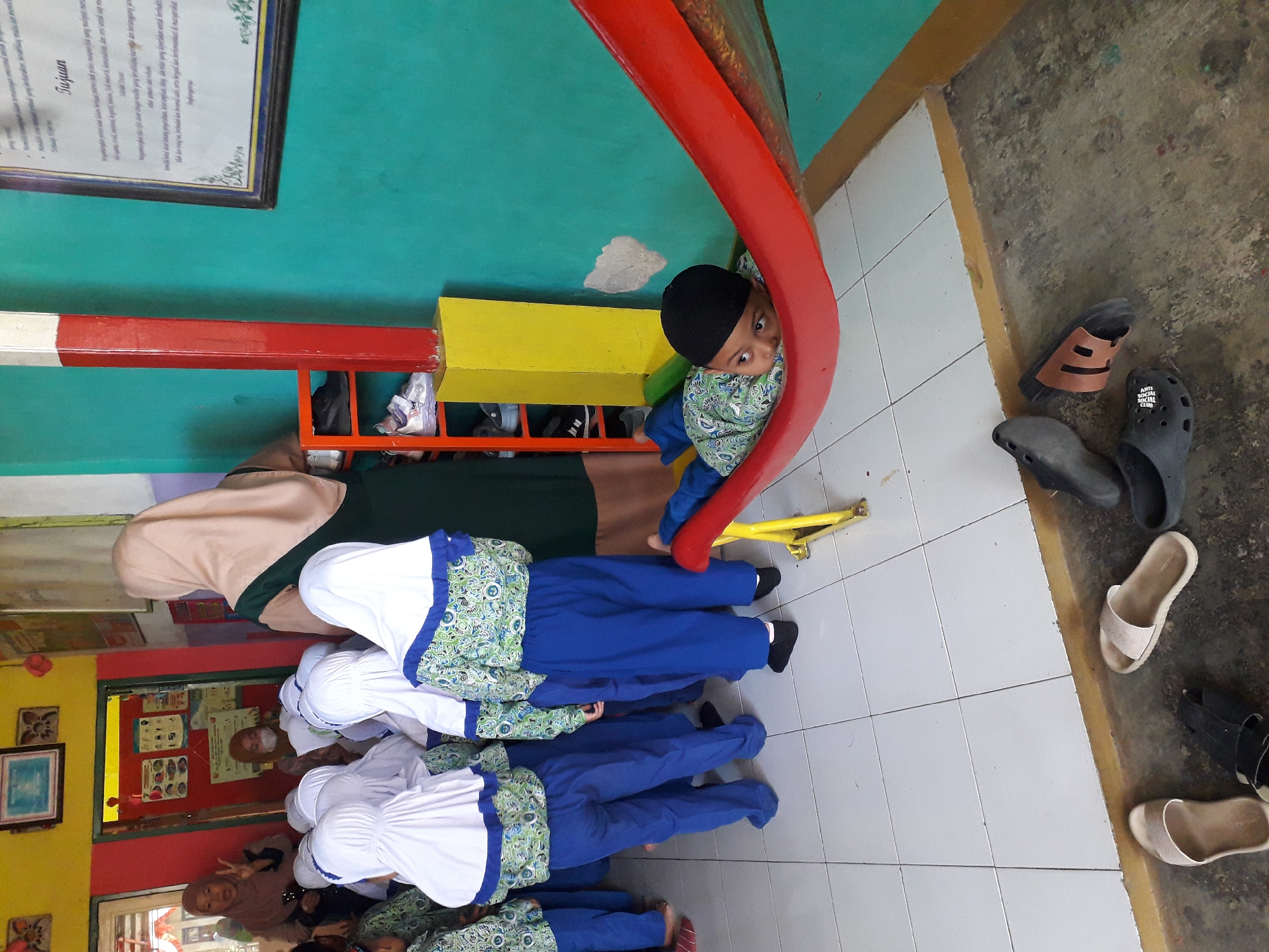 In Cankuang, Indonesia, a scene unfolds within what appears to be a classroom or community center. A group of children, dressed in matching blue pants and patterned tops with white headscarves, stand facing away from the camera. One adult, wearing a light pink headscarf and a dark green dress, stands behind them, facing towards a doorway. Through the doorway, another adult, wearing a lighter colored headscarf and a white mask, is partially visible, possibly interacting with the children.
To the right of the children, a young boy, wearing a black skullcap and a green and white patterned shirt with blue pants, is lying on a red hammock-like structure. He is looking towards the camera.
The environment features a brightly colored room with teal and yellow walls. A red and white striped pillar stands in the foreground. Shelving units with shoes are visible on the left. The floor is tiled in a light color and transitions to a rougher concrete floor where various pairs of sandals and flip-flops are scattered. A framed document with Indonesian text hangs on the teal wall. The lighting suggests an indoor setting, possibly during the daytime.