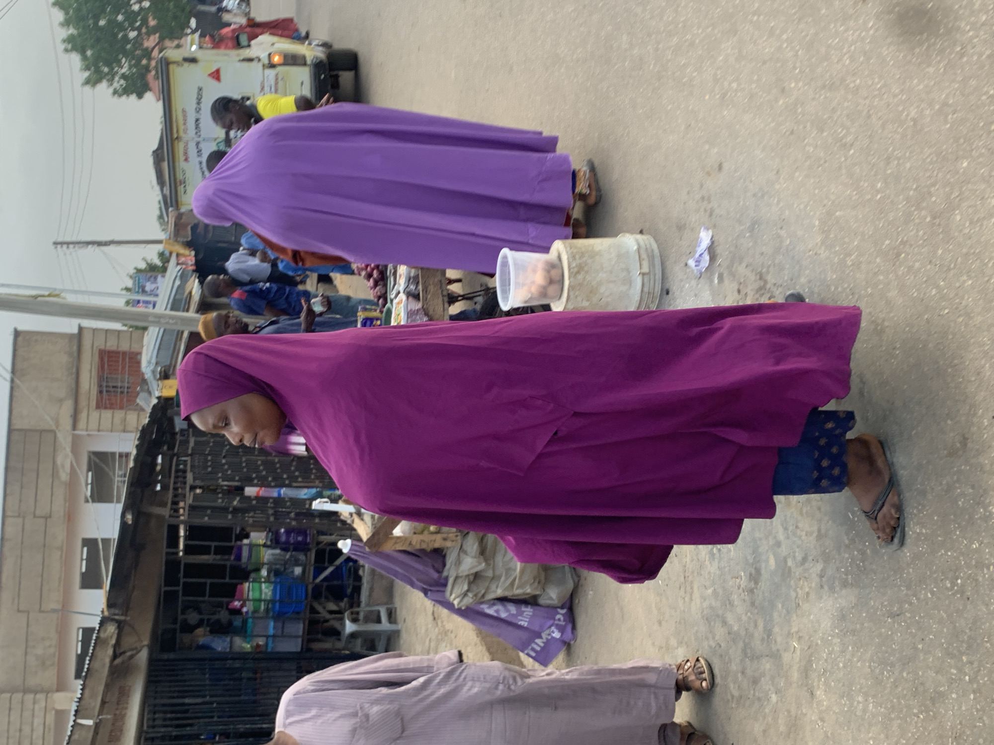 The image shows a street scene in Jos, Nigeria. Two women wearing brightly colored purple and lavender clothing are standing near a roadside stand selling food. One woman is wearing a purple head covering. The stand has containers of food products on it. There are other people, some of whom are standing near a vehicle, in the background. The scene is along a city street lined with buildings and shops.