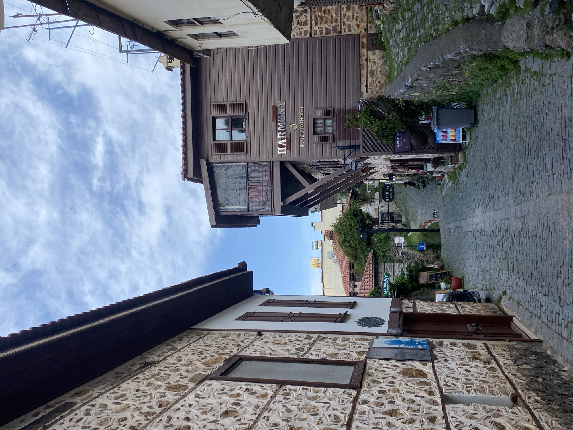 The image shows a narrow street in Alanya, Turkey, lined with buildings of traditional stone and wood construction. The sky is visible at the top of the image, with wispy clouds. On the left, a building has a stone facade with dark brown wooden trim. On the right, a wooden building with a sign that reads "Harmony Boutique Hotel" overlooks the street. The street is paved with cobblestones and slopes slightly uphill into the distance. Street signs and various vendor displays are visible on the right side of the street. No people are visible in the image.