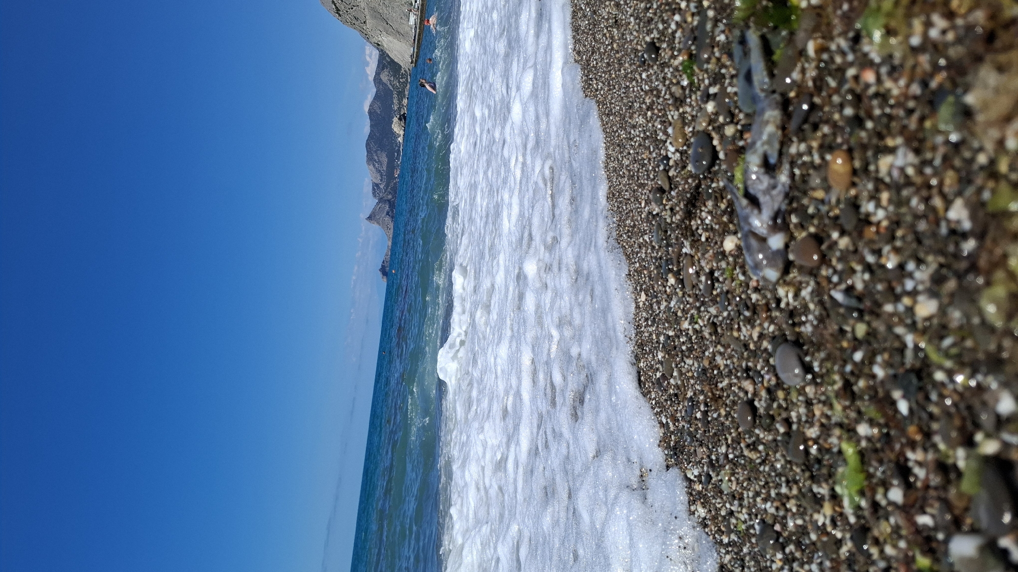 A close-up, low-angle shot of a pebbled beach on a sunny day. The foreground is dominated by the wet, multi-colored pebbles, with some scattered green seaweed and a small, gray sea creature lying on the shore. In the middle ground, white sea foam from a wave recedes from the shore. The background shows the turquoise ocean with a few people swimming and the rugged, mountainous coastline under a clear blue sky. The image is taken from a perspective near the water's edge, looking out towards the sea and mountains.