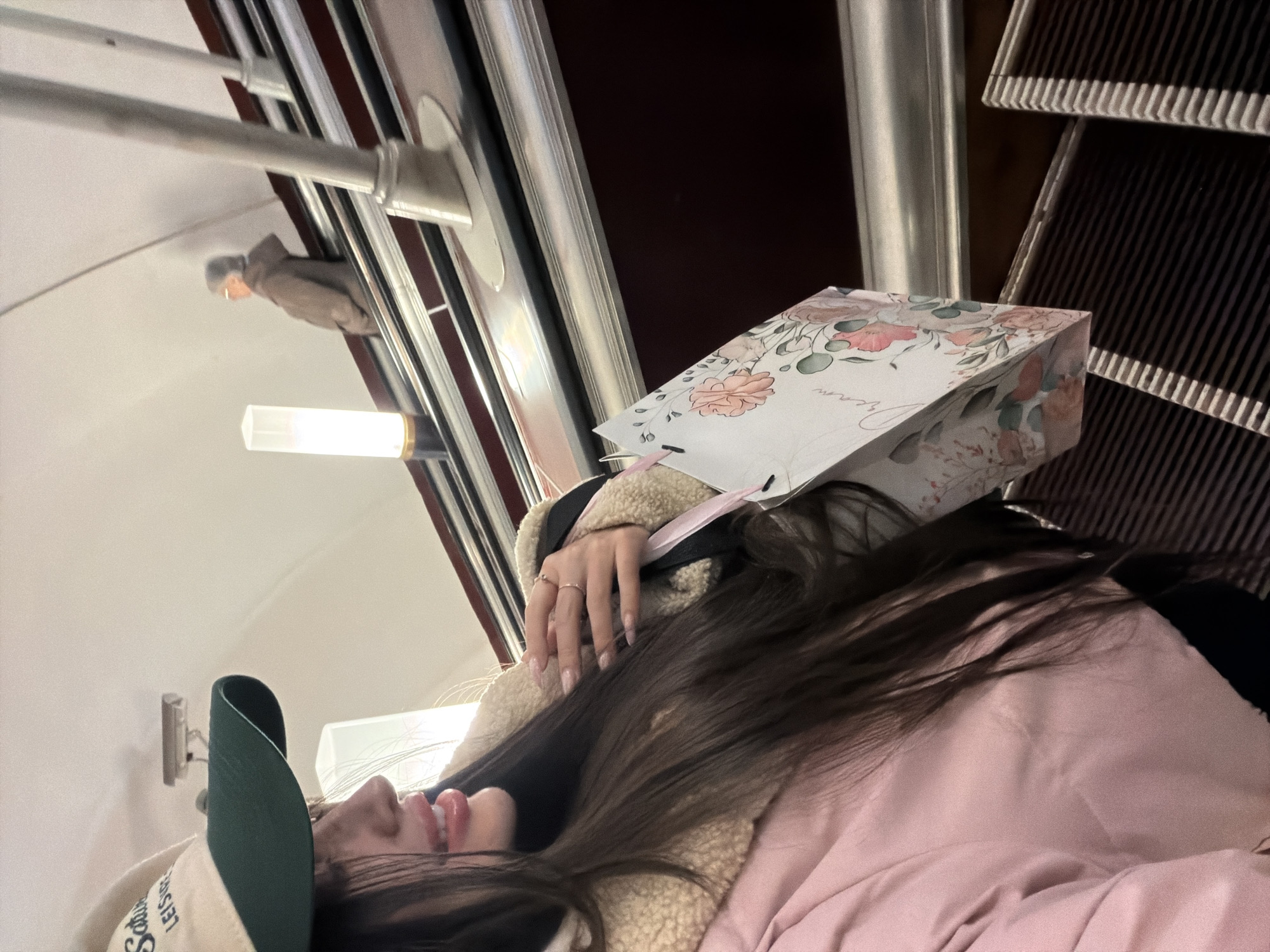 A young woman is on an escalator inside the Avtovo Metro station in Saint Petersburg, Russia. She wears a beige and green cap, a sherpa-lined jacket, and a pink jacket. A floral-patterned gift bag is positioned near her head. The escalator's moving stairs and metal railings are visible, as well as the station's wall and lighting fixtures. A statue is visible through the railing.