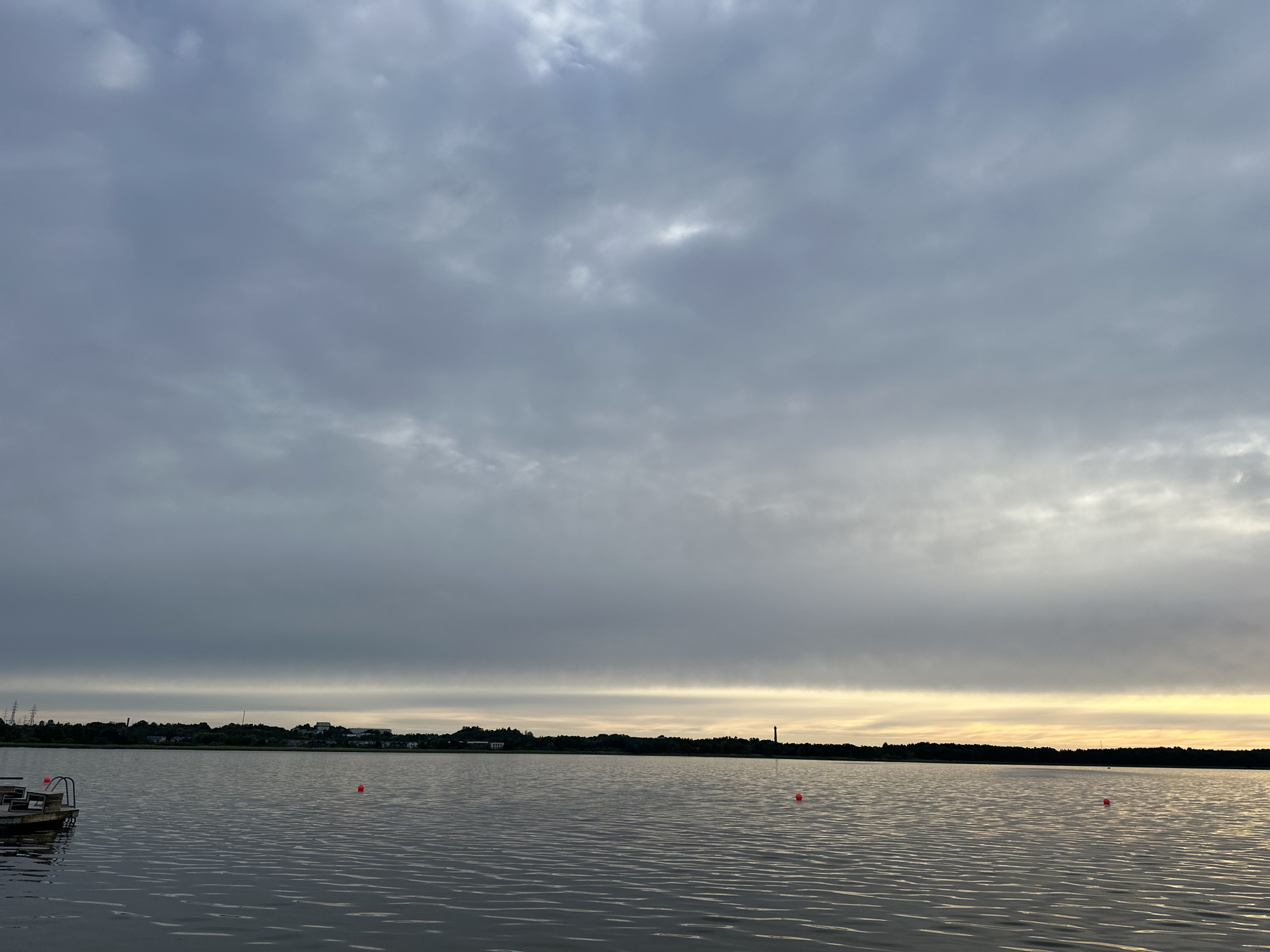 This image depicts a wide shot of a lake with a cloudy sky. The water is calm, reflecting the gray clouds overhead. On the left side of the image, a wooden dock with several chairs is visible, along with two orange buoys in the water. The far shore of the lake is lined with trees and scattered buildings, suggesting a developed area. The sky is dominated by thick, overcast clouds with a faint band of lighter, possibly sunlit, haze near the horizon. The overall impression is of a peaceful, possibly overcast, day at a lake. The location is likely a body of water within or on the outskirts of Tallinn, Estonia, given the presence of what appears to be a distant industrial structure or tower on the horizon.