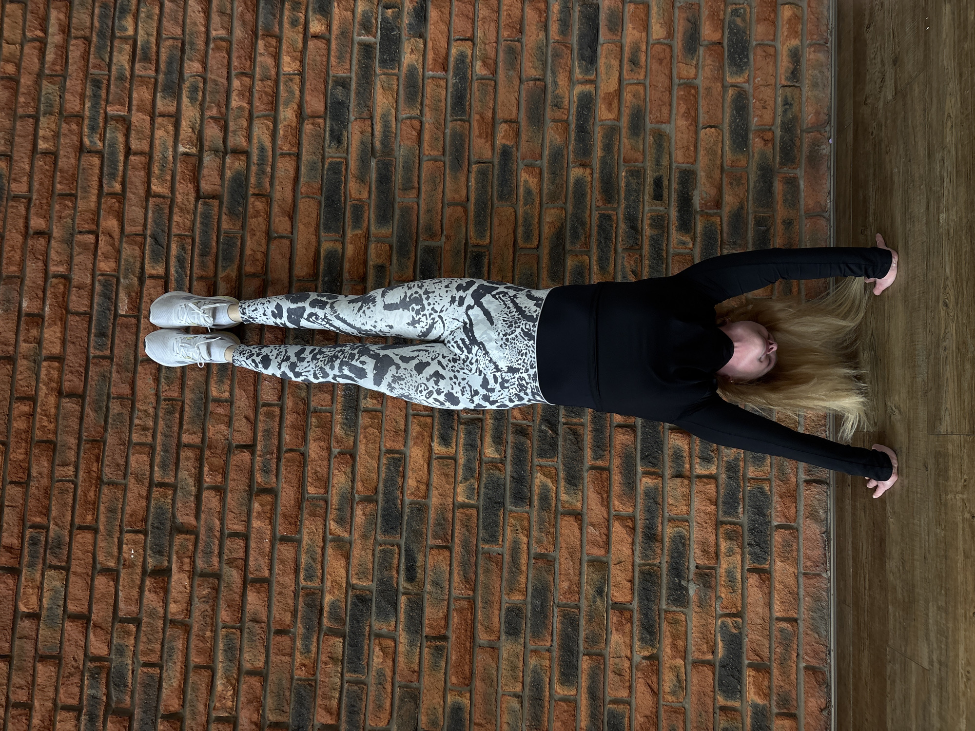 An overhead photograph captures a person lying flat on their back on a textured surface. The subject is likely female, with long blonde hair fanned out around their head. They are dressed in athletic wear, consisting of a black long-sleeved top, patterned grey and white leggings, and white athletic shoes.
The person's body is stretched straight and elongated, with arms extended upwards beyond their head and legs extended downwards. The pose suggests a static position, possibly part of a fitness routine, stretching, or a deliberate artistic composition.
The surface beneath the person is predominantly a brick floor or wall, featuring a pattern of reddish-brown and dark grey bricks. To the right side of the image, there is a distinct transition to a wooden floor, indicating an indoor setting where brickwork meets timber. The lighting appears even, without strong shadows, suggesting an artificial or diffused natural light source. No specific emotions are discernible, and there is no visible text in the image. The location context for this image is Chaikovsky, Russia.
