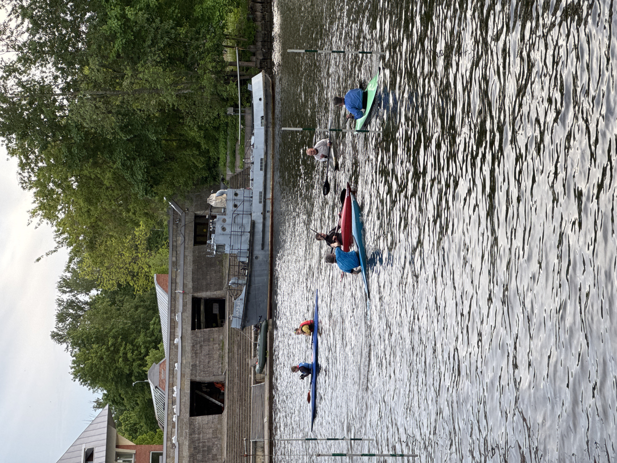 This image depicts a vibrant watersport scene unfolding on a waterway in Saint Petersburg, Russia, under bright daylight conditions. Several individuals are actively participating in what appears to be a canoeing or kayaking slalom training session or competition.
The foreground and middle ground of the image are dominated by the water, where multiple people are seated in various kayaks and canoes. A person in a vibrant green kayak is seen navigating between two white and green poles, which serve as slalom gates. Further back, another person is visible in a red canoe, while other participants in blue kayaks are positioned across the water, each equipped with a paddle. All individuals seem focused on their task, maneuvering their vessels through the course.
Along the left bank of the waterway stands a sturdy, multi-story building constructed from concrete or stone. Its utilitarian architecture suggests it could be a boathouse, a historical industrial facility, or a waterside structure. Moored directly alongside this building is a grey and white ship, possibly a patrol boat or a smaller naval vessel, distinguished by the numbers "33-05" clearly visible on its hull. A dark inflatable boat is also docked nearby.
Behind the building, the landscape is densely covered with lush green trees, hinting at a park or natural area adjacent to the urban waterway. The water's surface is textured with ripples, reflecting the clear sky and bright sunlight, indicating fair weather. The scene captures an energetic outdoor activity set against a backdrop of unique architecture and natural elements characteristic of Saint Petersburg's canals and rivers.