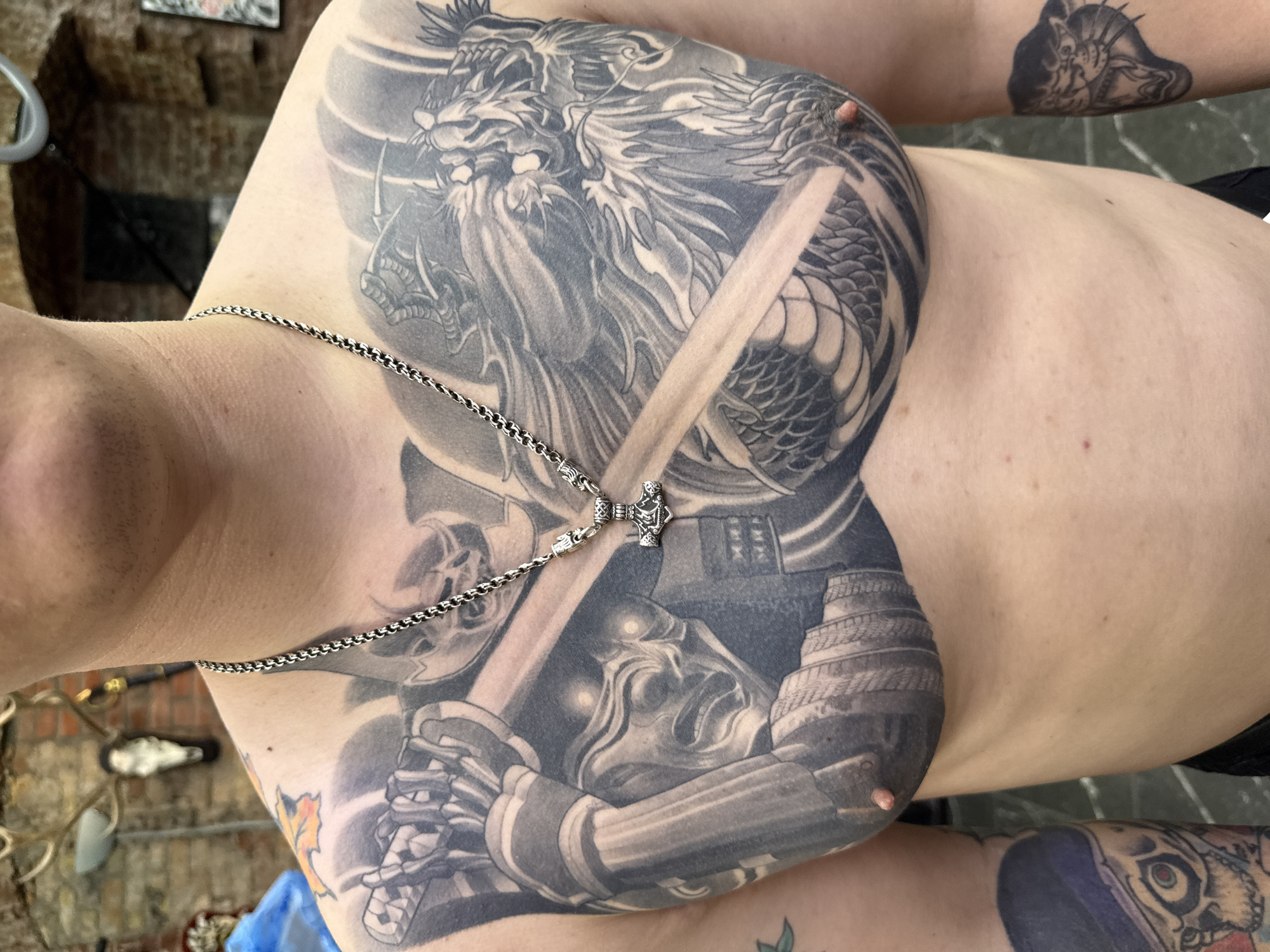 A shirtless person is shown from the neck down, with tattoos covering their chest and arms. A dragon tattoo covers the upper chest and right shoulder. A samurai figure with a sword covers the lower chest and left shoulder. The tattoos are black and gray. A silver chain necklace with a pendant hangs around the person's neck. The background is blurry but appears to be an outdoor setting with brick elements. The location is unknown.