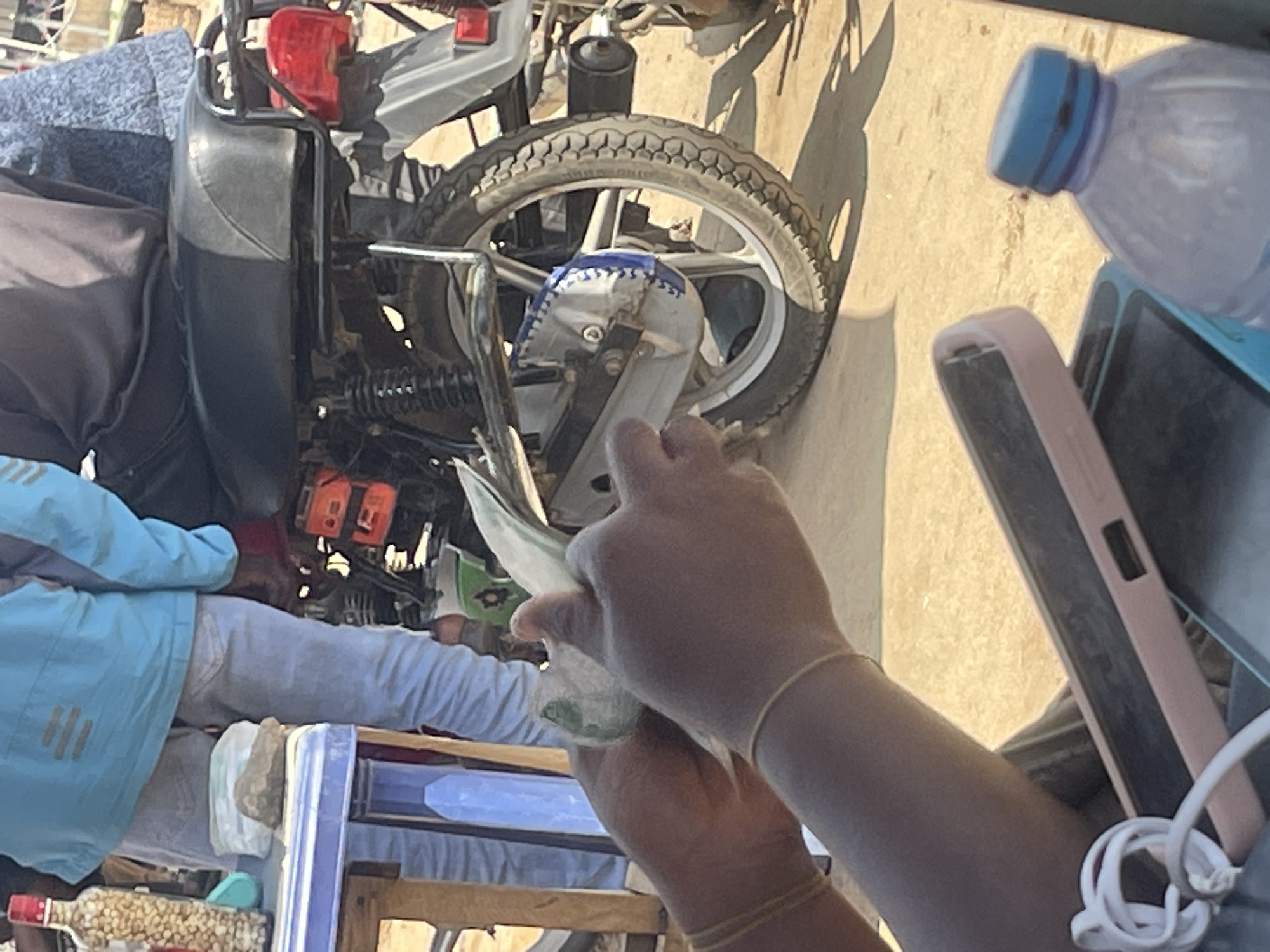 In the heart of Abuja, Nigeria, a scene unfolds with a blend of everyday activities and subtle indications of commerce. The image captures an individual's hands, clad in translucent gloves, meticulously handling what appears to be a bundle of cash, possibly in the process of counting or organizing it. The texture of the gloves and the faint green hues of the banknotes suggest an outdoor setting, with the bright sunlight casting sharp shadows, hinting at a daytime scene.
Behind the hands, the scene broadens to reveal elements of a bustling environment. A motorcycle is partially visible, its rear wheel and part of its frame indicating it's a mode of transport or a means of livelihood. Scattered around are various objects: a plastic bottle with a blue cap, likely containing water, a mobile phone in a pink case, and a tangle of white wires, possibly a charging cable. In the background, a person dressed in a light blue jacket and light-colored trousers is seated, adding to the human presence in the frame. The overall setting appears to be a roadside or an informal marketplace, common in urban Nigerian settings. The presence of the motorcycle, the cash, and the individual's focused action suggest a transaction or business-related activity. The sunlight and shadows indicate it is a clear day, and the overall mood appears practical and focused.