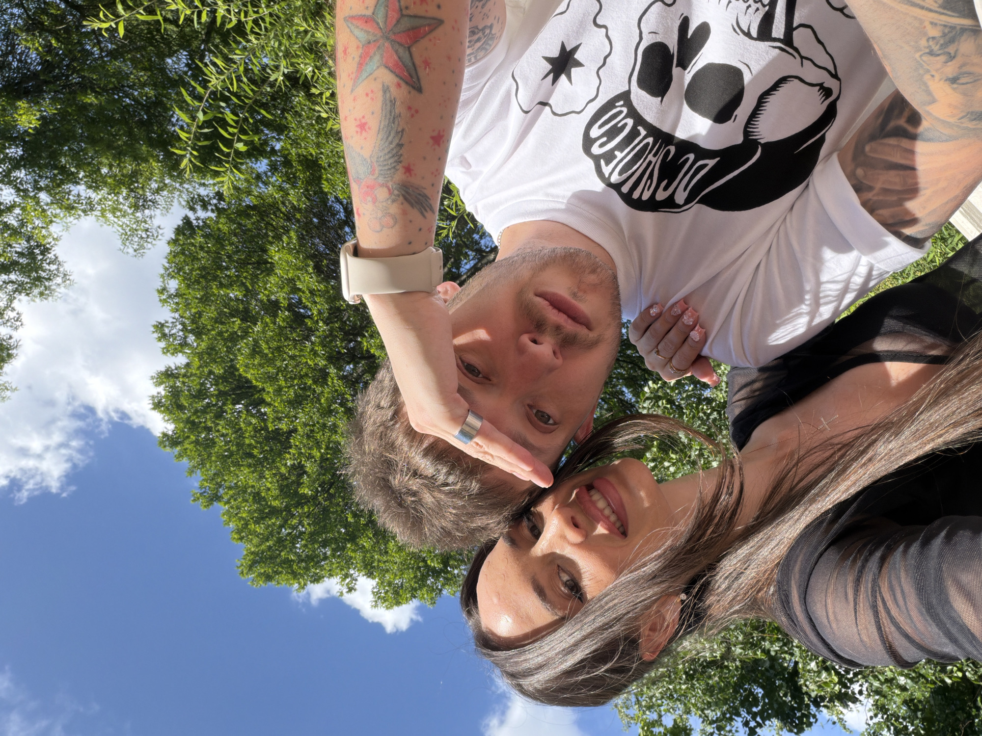 This image captures a close-up selfie of two individuals, a male and a female, in an outdoor setting in Saint Petersburg, Russia. The background features a clear blue sky with scattered white clouds and lush green tree foliage, indicating a bright, sunny day.
The female, positioned on the left and closer to the camera, has long dark brown hair parted in the middle. She is smiling broadly, looking directly at the camera, with visible teeth. She wears a sheer black long-sleeved top, a delicate gold chain with a cross pendant, and a small stud earring in her visible ear. Her right hand, displaying light pink manicured nails with a decorative pattern and a gold ring, rests on the male's shoulder.
The male, positioned to the right and slightly behind the female, has short light brown hair and light stubble on his face. He is also looking directly at the camera. His left arm is raised, with his hand, wearing a silver ring, partially covering his forehead, possibly shielding from the sun. A light-colored smartwatch is visible on his left wrist. His left forearm is extensively tattooed with a prominent red star, a red and black winged creature, and numerous small red stars. He wears a white t-shirt featuring a large black graphic print of a skull and the text "DC SHOE CO."
