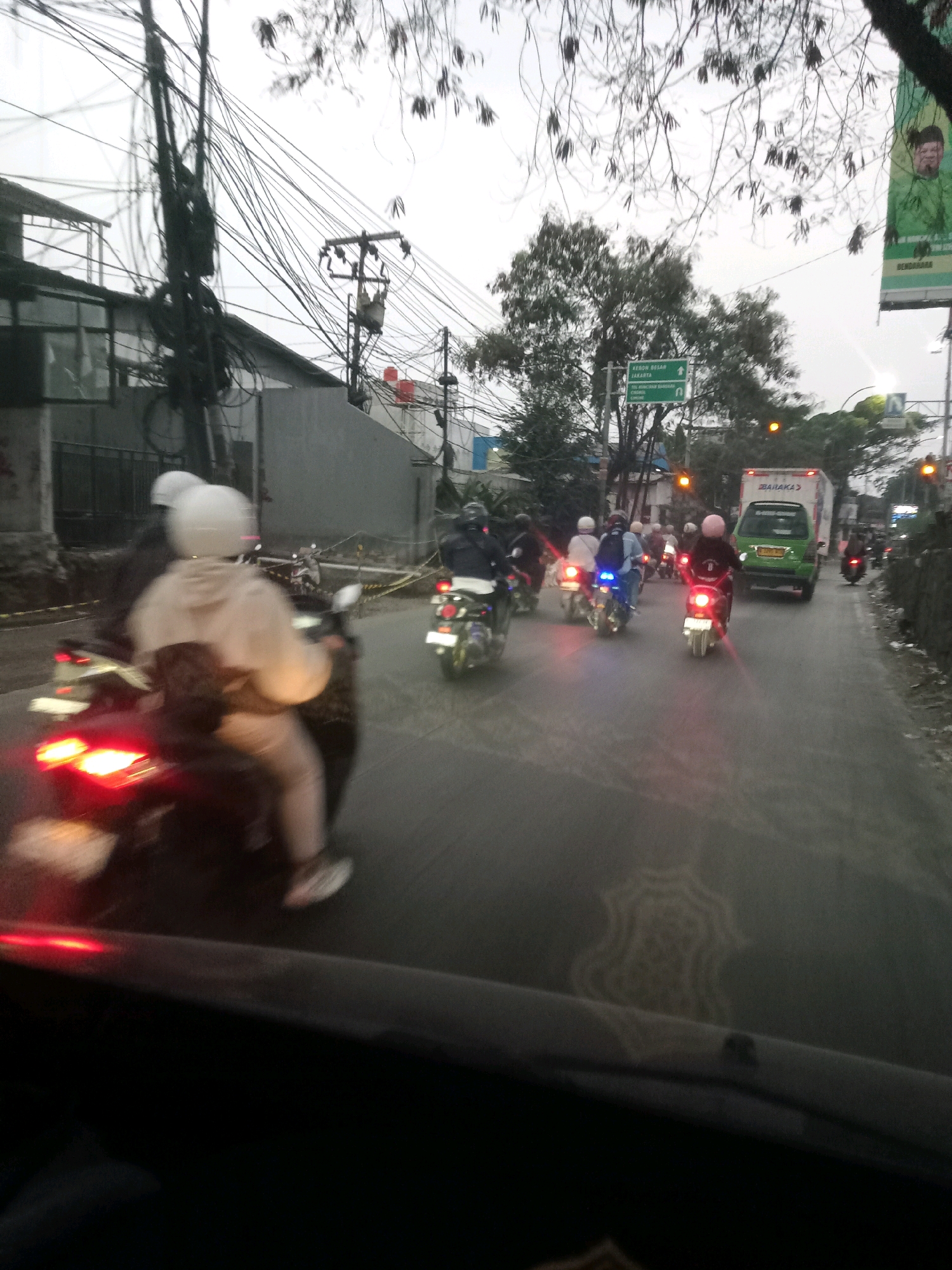 The image shows a street scene in Tangerang, Indonesia, likely during the day given the ambient light. A convoy of motorcycles is traveling along a paved road. Several motorcycles are visible, with riders wearing helmets and some carrying passengers. One motorcycle in the foreground has its brake lights illuminated. A green truck is also present in the traffic. The surrounding environment includes buildings, utility poles with numerous cables, trees, and a billboard. The road appears to be wet, suggesting recent rain. The overall impression is of moderate traffic flow in an urban setting.