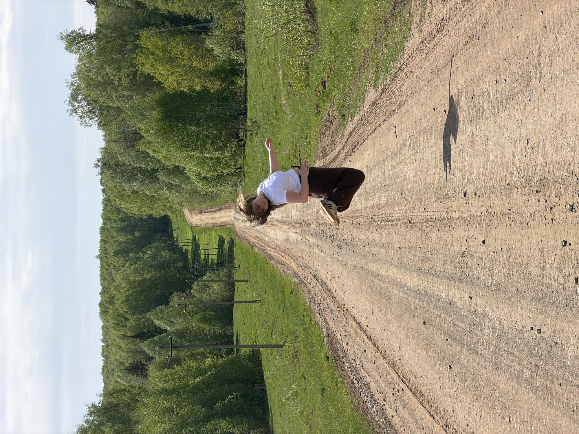The image depicts a dirt road curving through a grassy, wooded area. A person is posed mid-air on the road, seemingly suspended or jumping, casting a shadow. The location is implied as Ural'skoe, Russia, based on external context. Power lines run along the side of the road. The day is sunny.