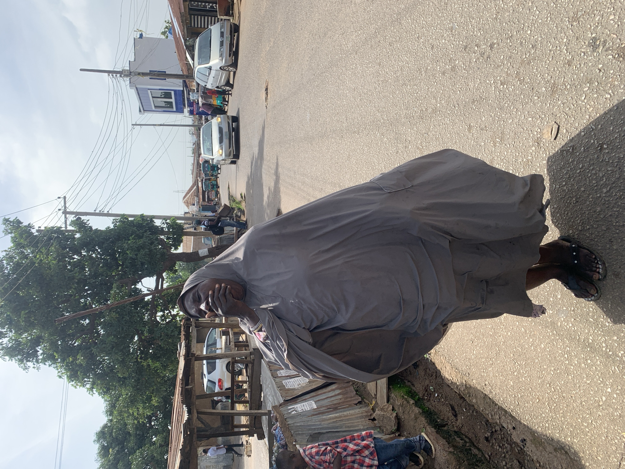 This media file captures a street scene in Jos, Nigeria. The primary subject is a person draped in a grey garment, who appears to be holding their face with their hand, possibly in a gesture of contemplation or distress. They are wearing sandals.
The scene is a roadside with asphalt paving and a dirt-lined gutter. Several vehicles are visible on the road, including cars and what appear to be three-wheeled auto-rickshaws, suggesting active traffic. In the background, buildings with varying architectural styles and storefronts are present, along with utility poles and a dense tree. The sky is overcast with clouds, indicating a possibly warm but not intensely sunny day. The overall impression is of a typical day on a busy street.
A child in a checkered shirt and jeans is lying on the ground near a corrugated metal structure, adding another human element to the scene. The lighting suggests it is daytime. There are no clearly discernible words or text that can be read from the image.
