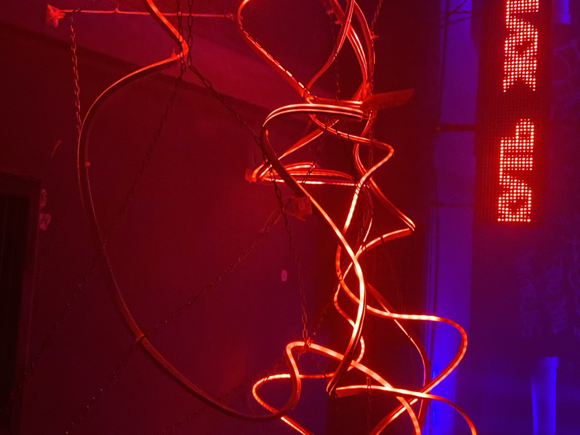 The image depicts an interior scene in Shebunino, Russia, likely a nightclub or bar, characterized by dim lighting and vibrant red and blue hues. A sculptural light fixture, suspended by chains, dominates the composition. This fixture features a large, circular frame and several lengths of flexible tubing illuminated with a bright, reddish-orange light. These tubes are arranged in a spiraling, chaotic manner within the frame. To the right of the light sculpture, a vertical LED sign displays pixelated text in red, though the exact words are partially obscured. The background is a dark, textured wall with hints of purple and blue lighting, suggesting a dimly lit, atmospheric environment.