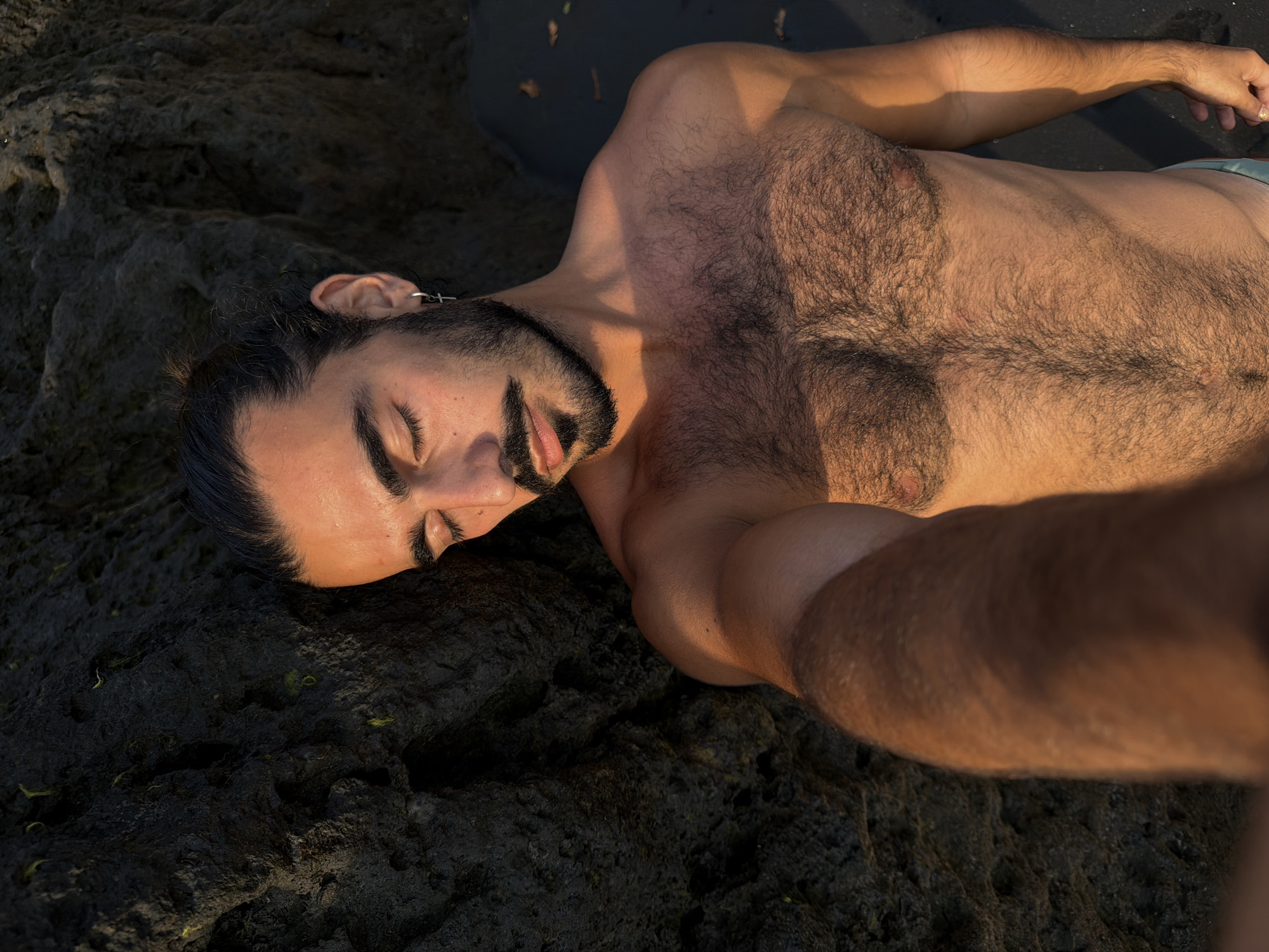 A man with dark hair pulled back in a bun and a beard reclines against a dark, rocky surface. He is shirtless, revealing a hairy chest and torso. His eyes are closed, and his head is tilted back. A small silver cross earring is visible in his left ear. The man appears to be outdoors, with a sandy or dark ground surface visible to his right. The lighting suggests it may be late afternoon or early morning, with warm sunlight highlighting his skin. The dark rocks and sand indicate a natural environment, possibly a beach or coastal area. The scene is set in Selemadeg, Indonesia.