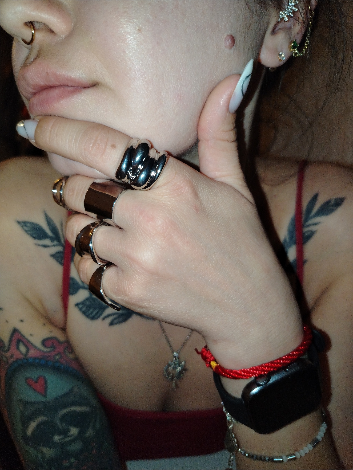 A person with pale skin is shown from the chest up, adorned with jewelry and tattoos. A gold nose ring is visible in the left nostril, and multiple earrings decorate the right ear. The person's left hand is held near their face, and several large silver rings are worn on the fingers. The person's nails are long and painted with a white design. A tattoo featuring a raccoon with a heart is visible on the left upper arm, and there are leaf tattoos on the upper chest/shoulders. A silver necklace with a pendant is visible on the chest. On the right wrist, a black smartwatch is worn along with a red braided bracelet and a beaded silver bracelet. The person is wearing a red top. The environment is not identifiable in terms of geographic location and is not definitively linked to the city of Noviy, Russia.