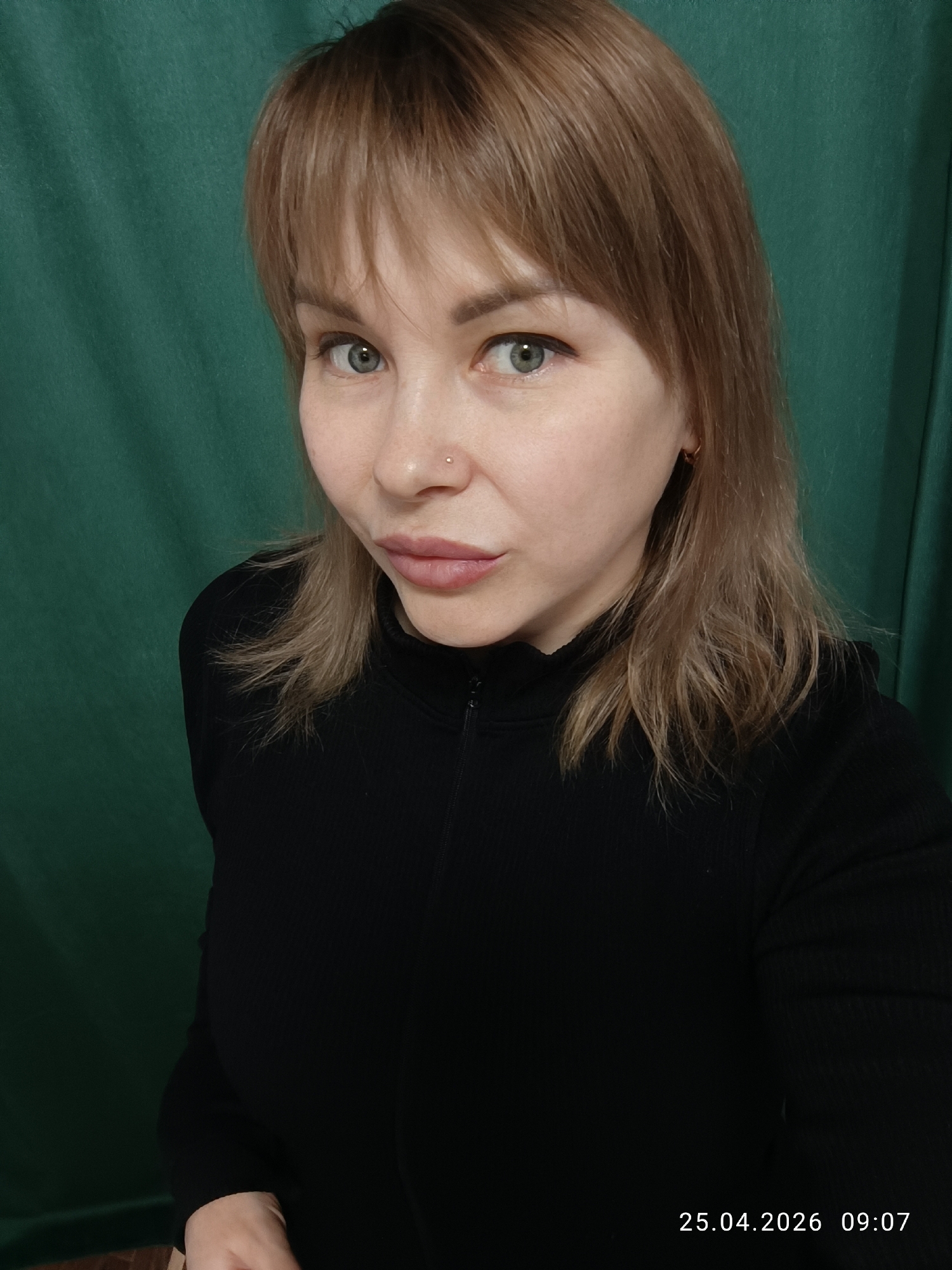 A woman with light brown hair and bangs is looking at the camera. She has fair skin, green eyes, and a nose piercing. She is wearing a black, long-sleeved shirt or sweater with a small zipper at the neck. The background is a solid green color, possibly a curtain or wall. The date "25.04.2026" and time "09:07" are printed in white at the bottom right corner of the image. There is no location or reference to the city of Chaikovsky, Russia.