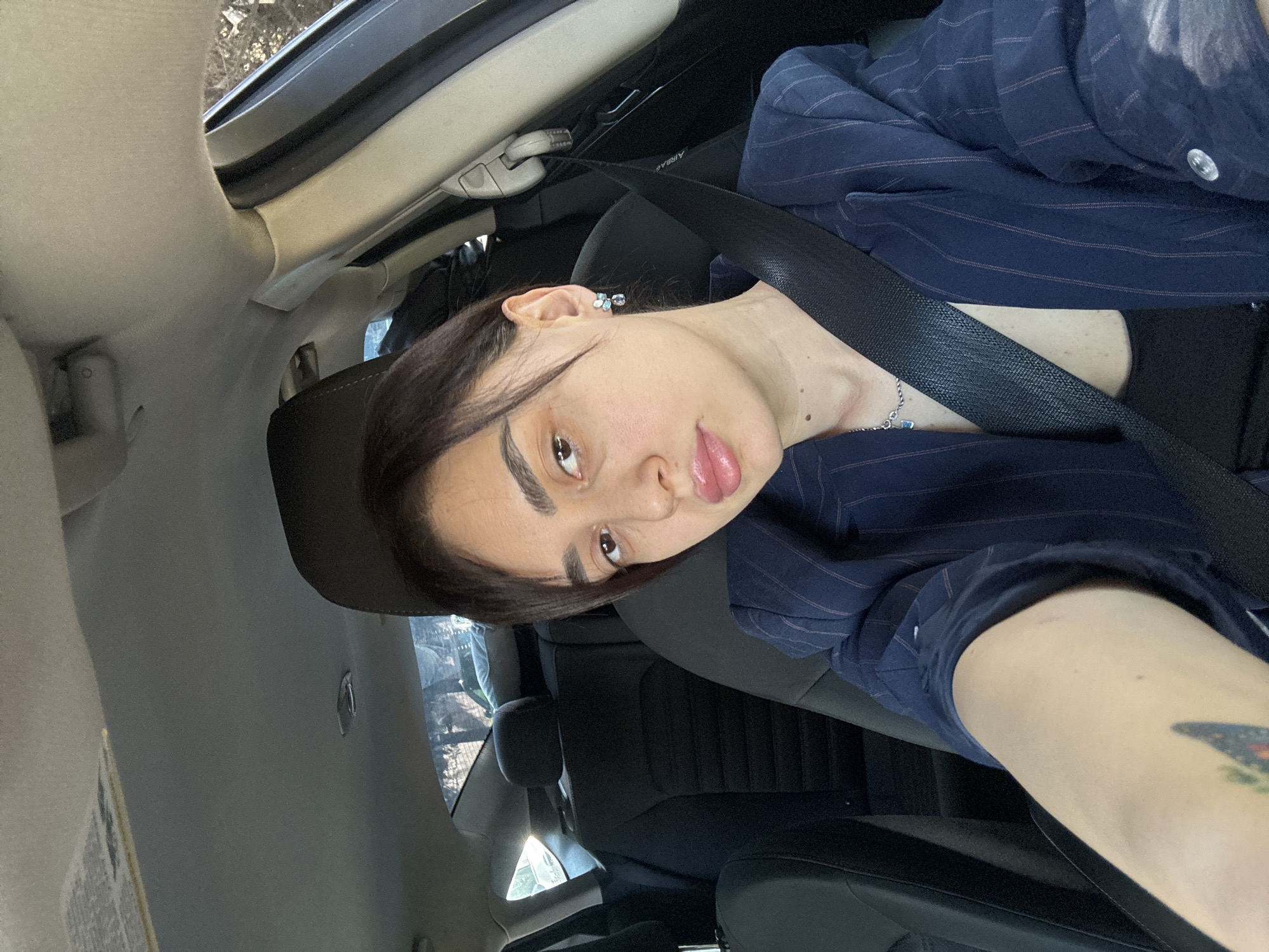 A woman is in the driver's seat of a car. She is wearing a navy pinstripe blazer over a black top, and a seatbelt is fastened across her. Her hair is dark and pulled back from her face. The interior of the car is visible. The location is not discernible from the image.