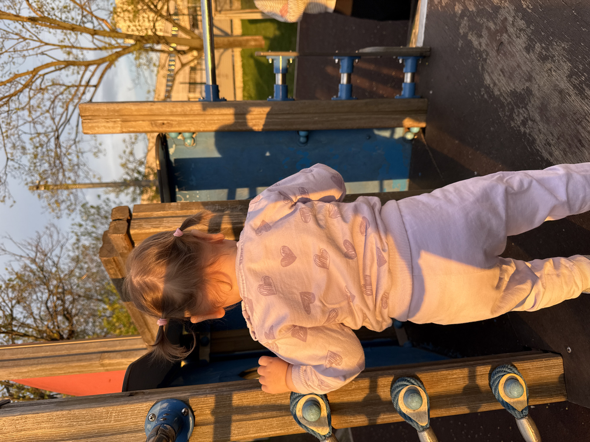 A young child with light hair styled in pigtails, wearing a light purple heart-patterned sweatshirt and matching pants, is crawling on a wooden structure with blue and silver metal components. The structure appears to be part of a playground. The lighting suggests it is late afternoon or early evening, with warm sunlight casting shadows. In the background, there are trees and buildings, suggesting an urban or suburban park setting. There is no information that suggests the location is in Moscow, Russia.
