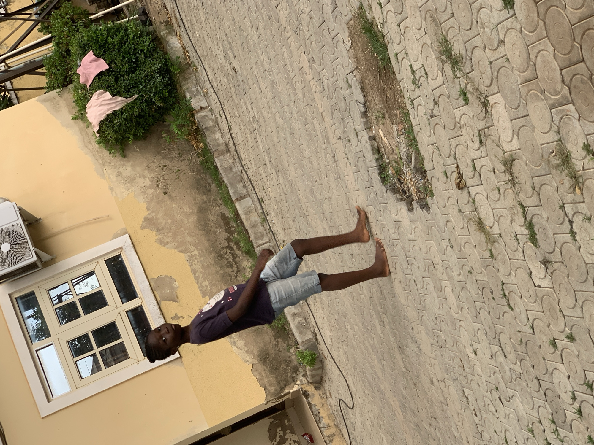 A young Black boy, barefoot and wearing a dark purple t-shirt and light blue denim shorts, is captured in a full-body, slightly angled shot as he walks on a paved courtyard. The boy's gaze is directed towards the camera with a neutral expression. To his left, a pale yellow building features a large window with multiple panes and an air conditioning unit mounted above it. In the background, to the right of the boy, a dense green bush is adorned with two pieces of pink fabric draped over its branches, possibly laundry drying in the sun. The ground beneath the boy is made of interlocking concrete pavers, with patches of dirt and grass visible in the lower right quadrant, indicating a somewhat unkempt outdoor environment. The overall scene suggests a residential setting, possibly in the Garki district of Abuja, Nigeria, given the architectural style and the presence of an air conditioning unit.