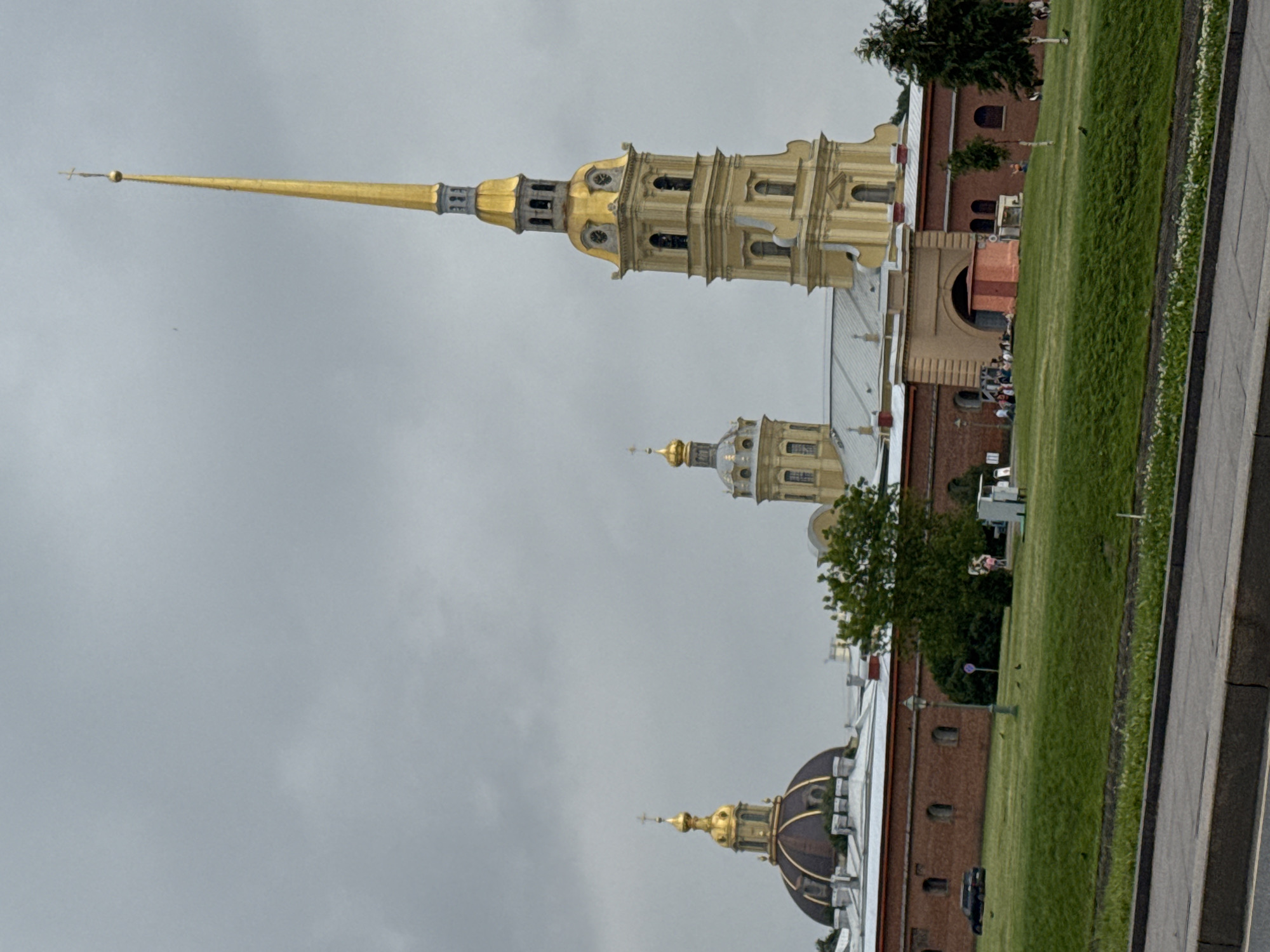 The image shows the Peter and Paul Fortress in Saint Petersburg, Russia. The fortress is a historic citadel that includes the Peter and Paul Cathedral, a prominent landmark with a tall, golden spire. Several other buildings with golden domes and spires are visible, set against a gray, overcast sky. The fortress walls are made of red brick, and a large grassy area with trees and scattered people is in front of them. A road with a sidewalk is in the foreground.
