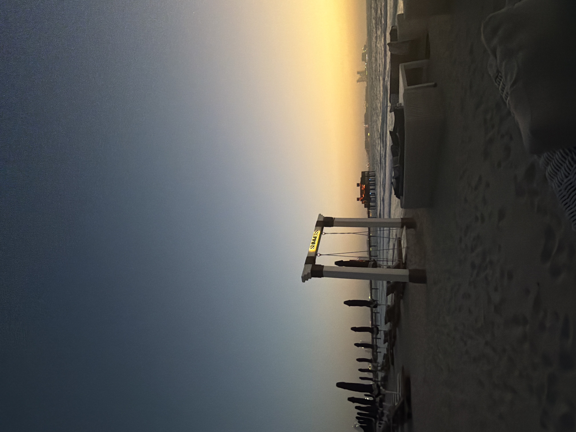 The image depicts a beach scene at sunset in Dubai, United Arab Emirates. The sky transitions from deep blue to a golden hue near the horizon. A row of closed black umbrellas and beach chairs lines the sandy beach. A white structure, possibly a swing set or decorative archway, with the word "SUMMERS" illuminated on its top is visible. In the distance, a pier with lights and the city skyline are silhouetted against the setting sun. The foreground shows sand and part of a white structure and a person.