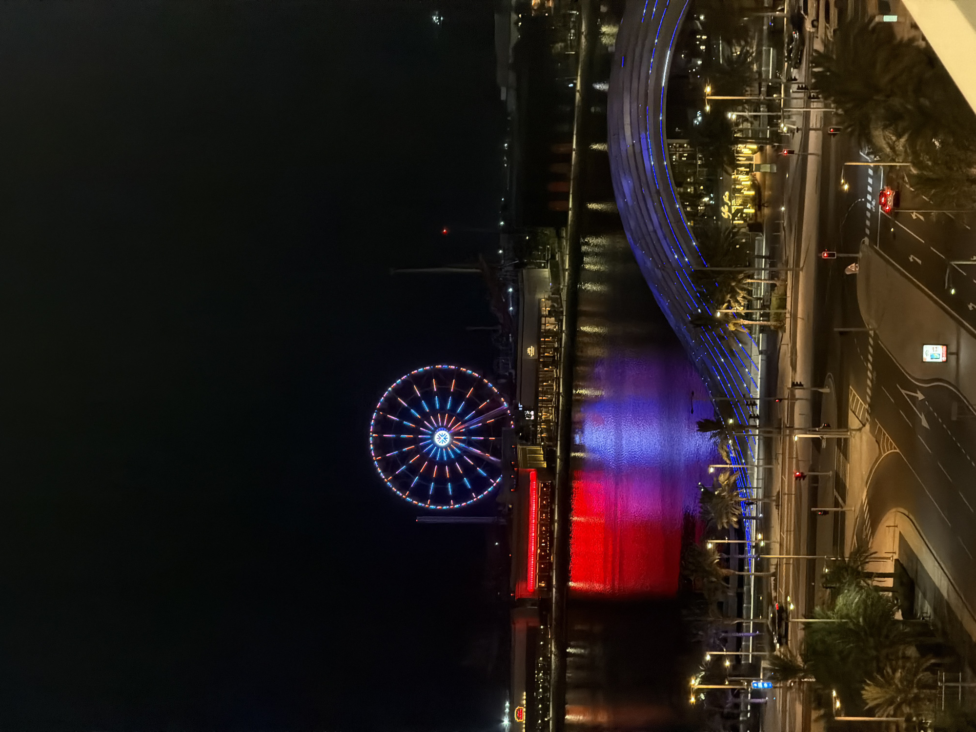 An elevated, full-frame, night-time shot captures a vibrant waterfront scene. Dominating the mid-ground is a large Ferris wheel, illuminated with a radial pattern of red, blue, and white lights. Below it, a building complex runs along the water's edge, with some windows lit warmly, and red and blue lights reflecting prominently on the dark water. In the foreground, a modern, arched bridge with blue lights crosses over a street. The street below is paved, with lane markings and traffic lights visible. A few cars are present on the street, and palm trees line the sidewalks and some areas of the waterfront. The sky is completely dark.