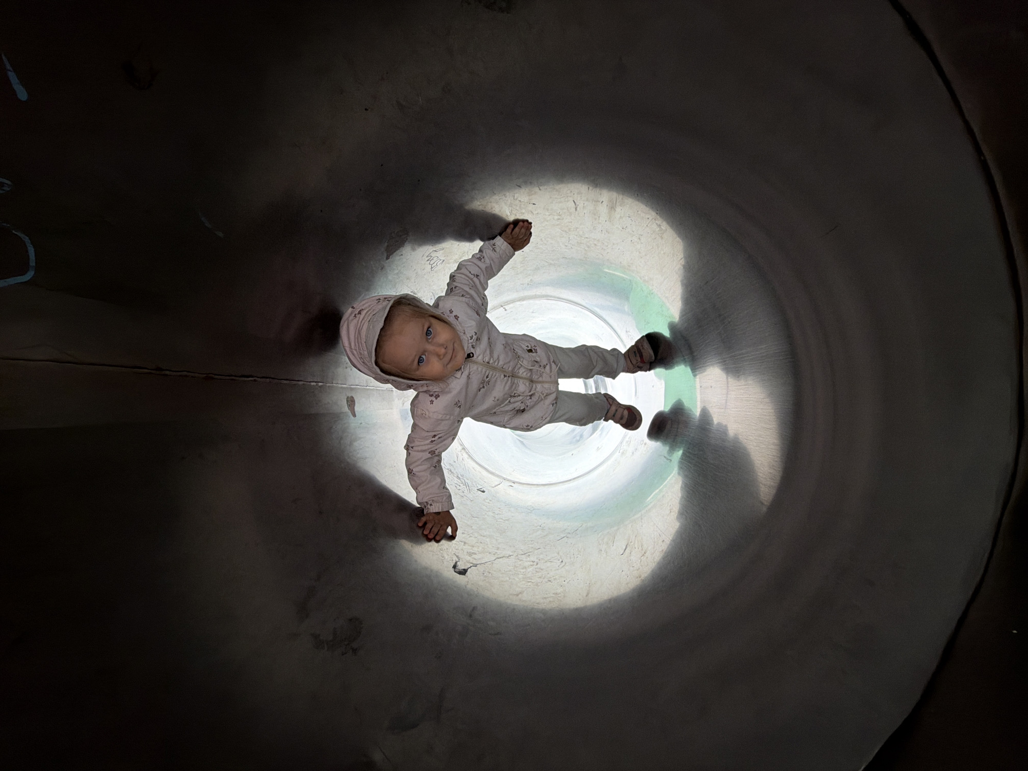 A young child, appearing to be a toddler, is shown from a high angle view, lying down inside a curved, metallic tube, likely a playground slide. The child is wearing a light-colored hooded jacket with a floral pattern and light-colored pants, along with pink shoes. Their head is turned towards the camera, and they have blue eyes and fair hair. The child's body is oriented diagonally within the tube, with their arms and legs spread slightly.
The tube itself is dark on the outer edges, gradually becoming lighter towards the center, suggesting it's a tunnel with light coming from the far end. The metallic surface of the tube shows some texture and faint markings. The lighting creates a spotlight effect on the child and the area immediately around them. The scene appears to be indoors or in a shaded outdoor area, given the contained environment of the tube. There is no visible text in the image. The child's expression is neutral or slightly curious. The overall impression is a playful moment captured within a children's play structure.