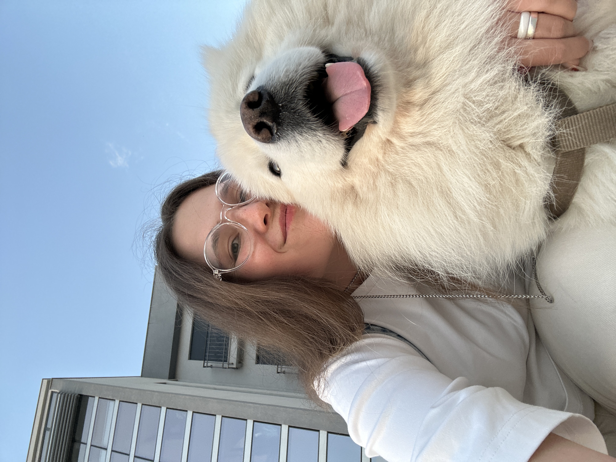 A medium shot angled upward shows a woman holding a Samoyed dog close to her face. The woman has brown hair, fair skin, and is wearing round-rimmed glasses. She is wearing a white long-sleeved shirt and a thin silver chain. Her face is turned slightly towards the right. The Samoyed is pure white with its tongue sticking out. It wears a brown harness. A silver ring is visible on the woman’s hand holding the dog. Behind the woman and the dog, a modern building with large reflective windows is partially visible. The sky is light blue. The location is Šušanj, Montenegro.