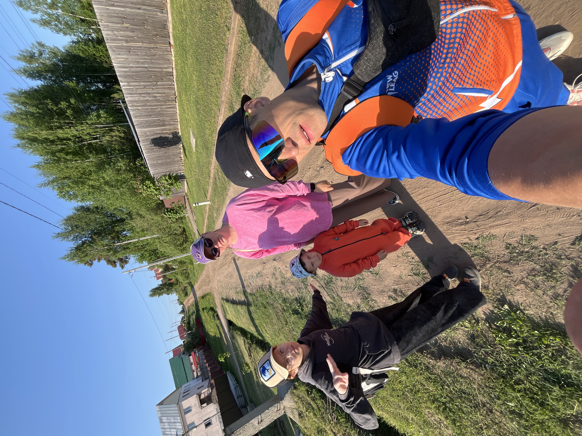 The image shows a group of four people lying on the ground, likely posing for a selfie taken by one of them. The setting appears to be a rural or suburban area in the city of Chaikovsky, Russia. The background includes a wooden fence, trees, and some buildings including a house with a grey roof. There is also green grass and a dirt path.
The person closest to the camera is wearing a blue and orange sports jacket, a black cap, and reflective sunglasses. Next to this person is another person wearing a pink shirt and a purple cap. To the right of the pink-clad person is a child in an orange snowsuit and blue hat. The person furthest from the camera is wearing a dark sweatshirt, dark pants, and a baseball cap. They are making a peace sign with their fingers.