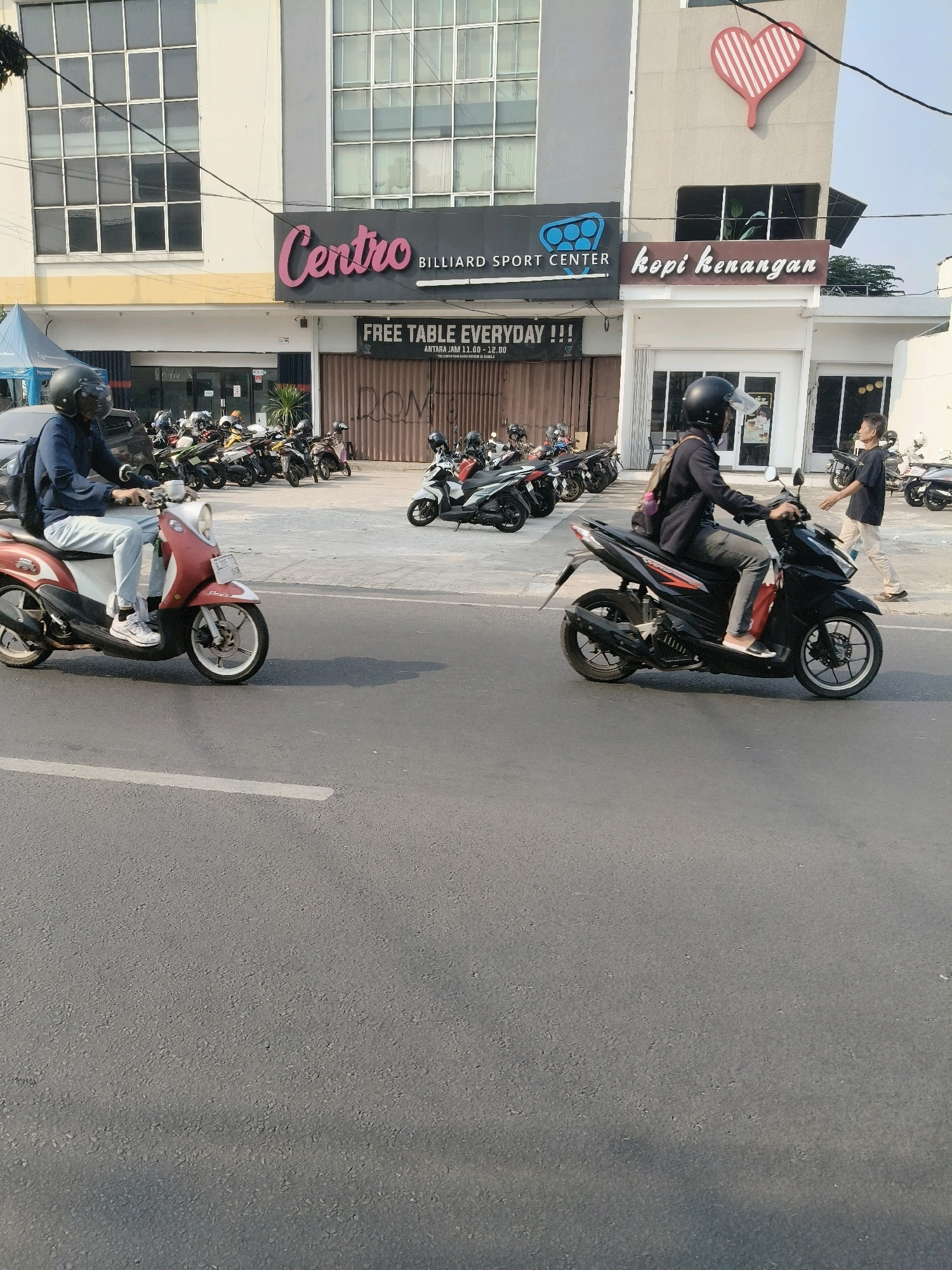 In South Tangerang, Indonesia, on a sunny day, two individuals are riding motorcycles on a paved road. The motorcycle on the left is red and white, and the rider wears a blue long-sleeved shirt, light blue jeans, white sneakers, and a black helmet. The motorcycle on the right is black, and the rider wears a dark jacket, grey pants, and a black helmet. The background features a building with signs for "Centro Billiard Sport Center" and "Kopi Kenangan," indicating a commercial area. Several other parked motorcycles are visible in front of the building, as well as a person walking in the background. The road surface is asphalt with visible lane markings.