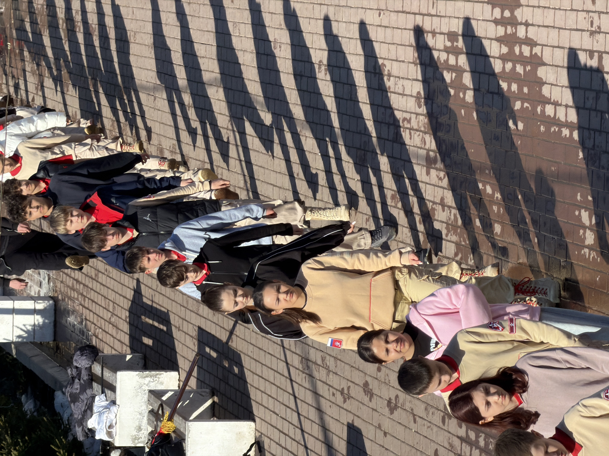 A group of people, mostly young adults, are lying in a line on a paved surface. The location appears to be outdoors, based on the presence of sunlight and shadows. The shadows cast are from overhead structures, possibly trees or building elements. A set of concrete steps and a cleaning tool (a mop or brush) are visible to the left side of the image. Some of the people are wearing clothing with red accents or patches, some appearing like Russia-themed emblems. The setting appears to be Izhevsk, Russia based on the emblems. The people are lying in a line, relatively close together, with their heads aligned in the same direction. The paved surface appears to be a public walkway or plaza.
