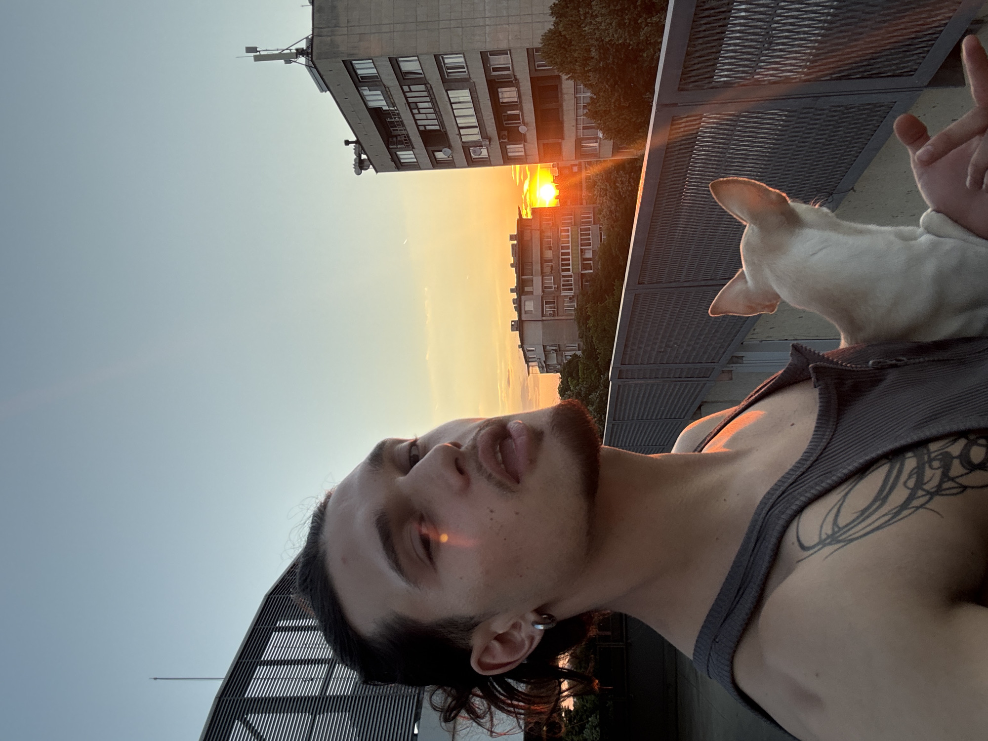 A young man with dark hair and a goatee is taking a selfie on a balcony. He is wearing a grey tank top and has a tattoo on his arm. A small white dog is perched on his shoulder, looking away from the camera. The sun is setting behind apartment buildings, casting a warm glow. The sky is a clear, pale blue. The man appears relaxed and content. The scene suggests a peaceful evening in a city. The image was likely taken in Sofia, Bulgaria, given the architectural style of the buildings, which are common in the city. The time of day is sunset.