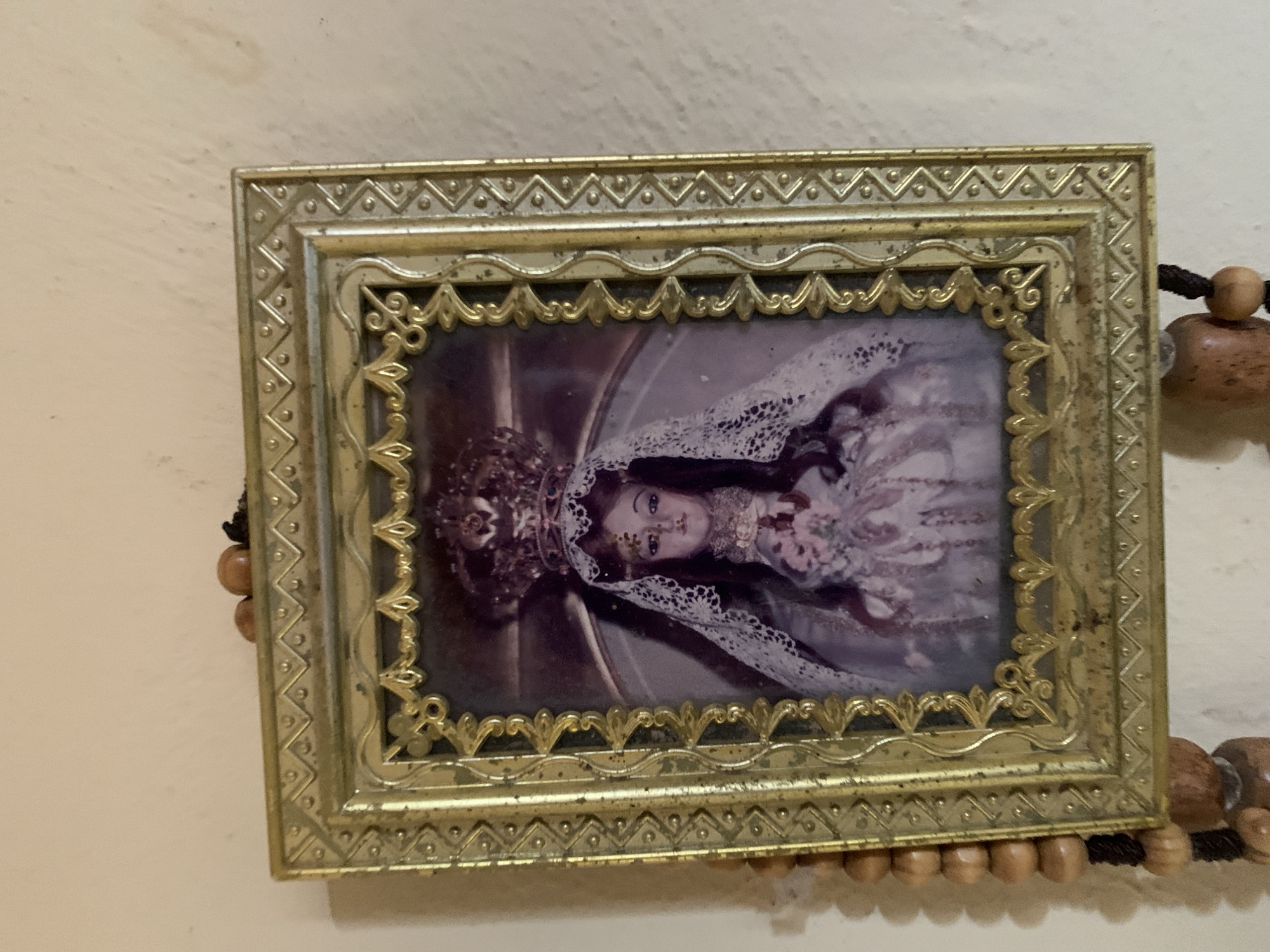 A gold-colored, ornate, rectangular picture frame hangs on a light beige or off-white wall. Inside the frame is a photograph of a woman with fair skin, wearing a white lace veil and crown. She appears to be holding flowers or some other adornment in her hands. The image and frame are mounted on a rosary with tan, wooden beads. The beads are strung on black rope.
The location is unknown and cannot be confirmed by the provided image.