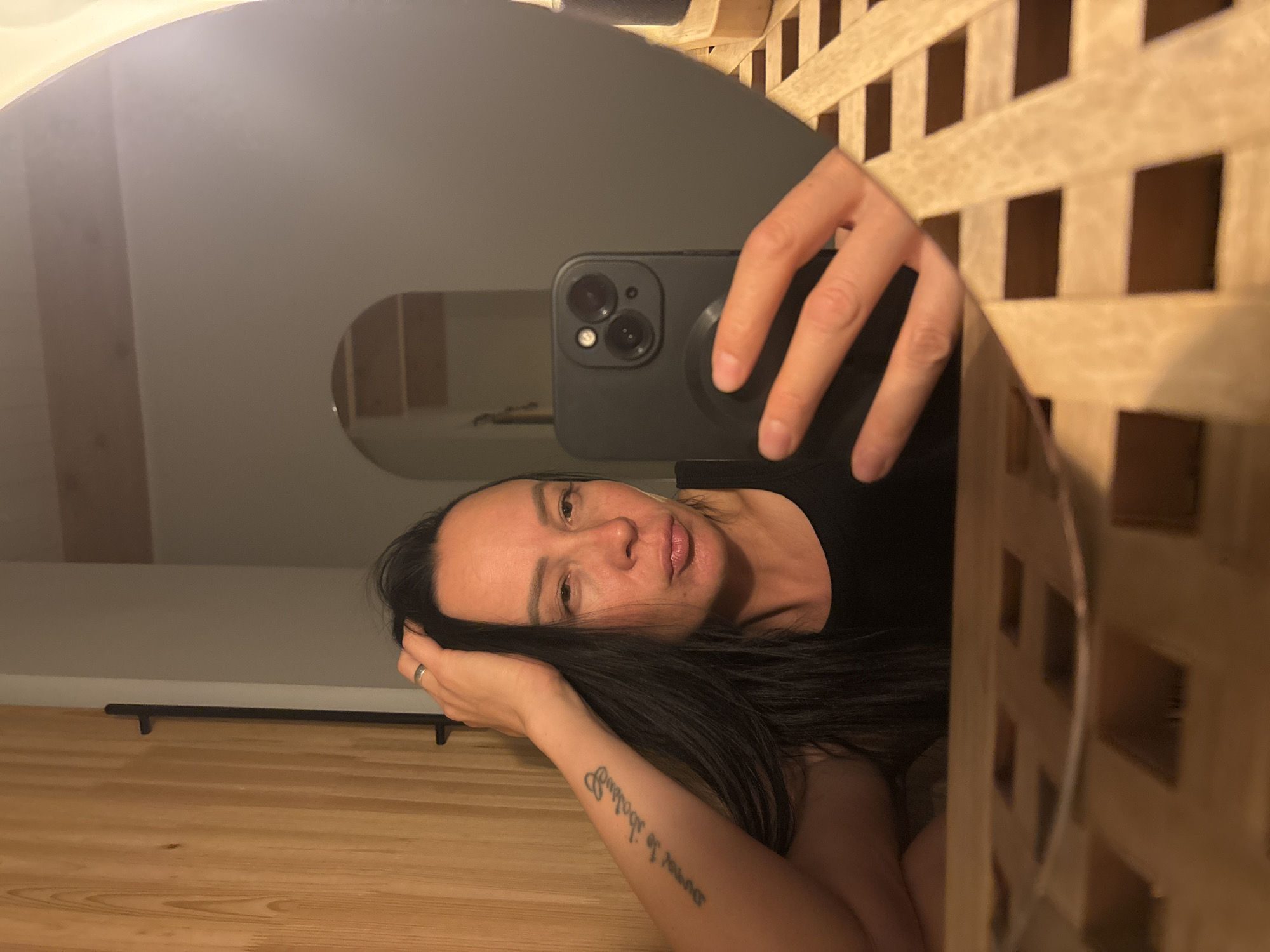 A woman is reflected in an oval mirror. She holds a smartphone to take a selfie. She has long dark hair, and a tattoo is visible on her inner left forearm. The mirror is framed by a light-colored, lattice-like wooden structure. Behind the woman's reflection is a neutral-toned room with an arched doorway. The reflection also shows a wooden floor with a dark bar running horizontally across it. There is no indication of location in the city of Chaikovsky, Russia, or any other specific location.